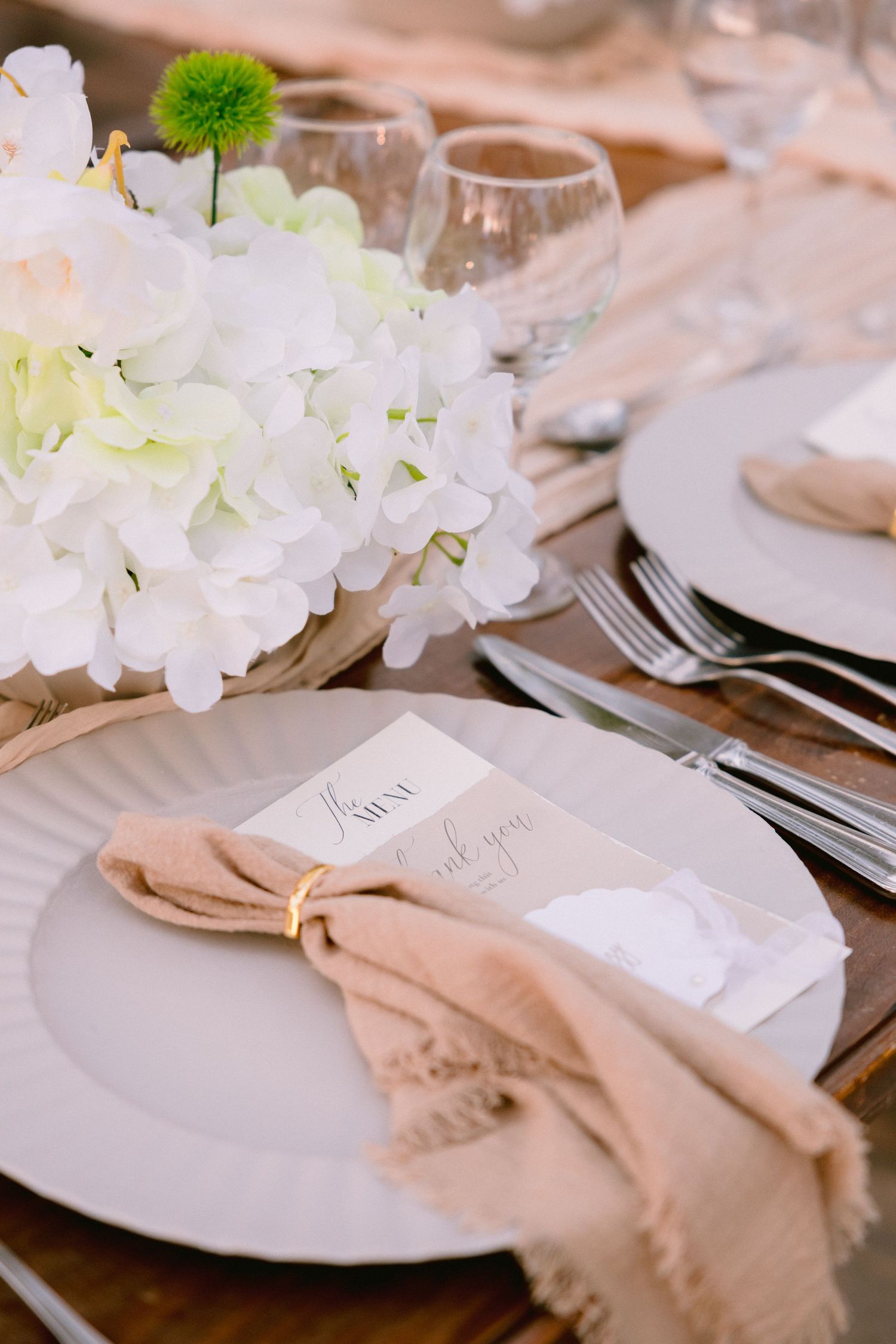 Elegant wedding table setting with white hydrangeas, neutral linen napkin, menu card, and gold ring on gray charger plate.