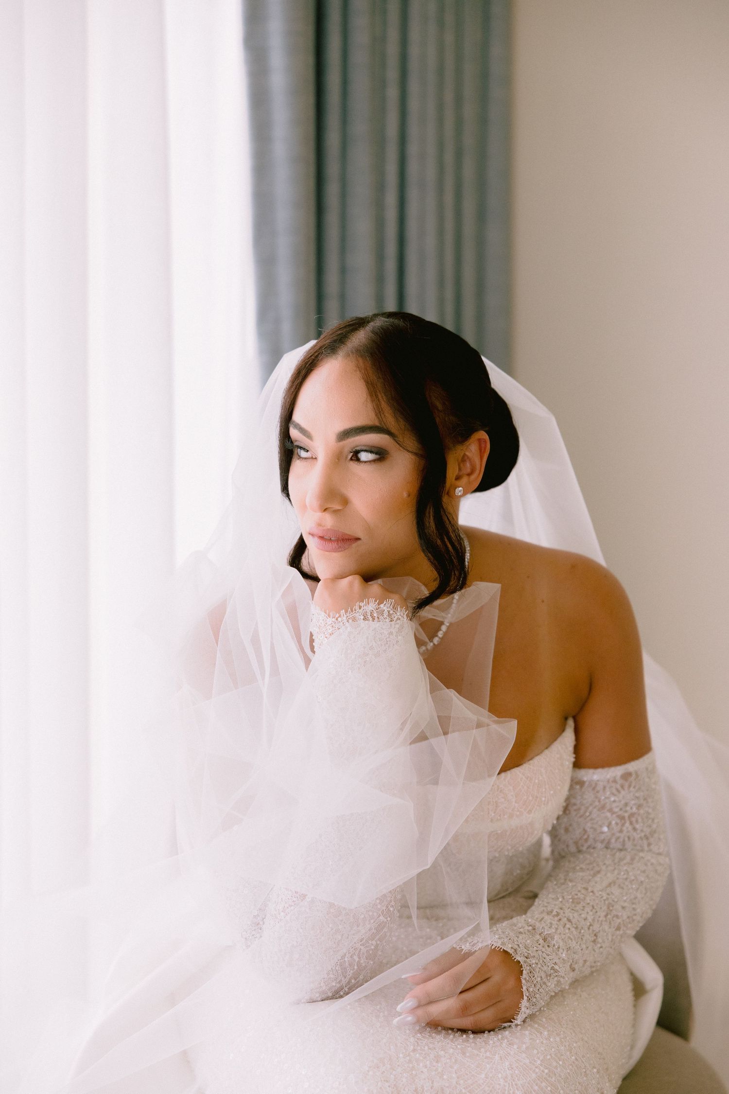 A bride in an elegant white dress and veil poses playfully near white curtains.