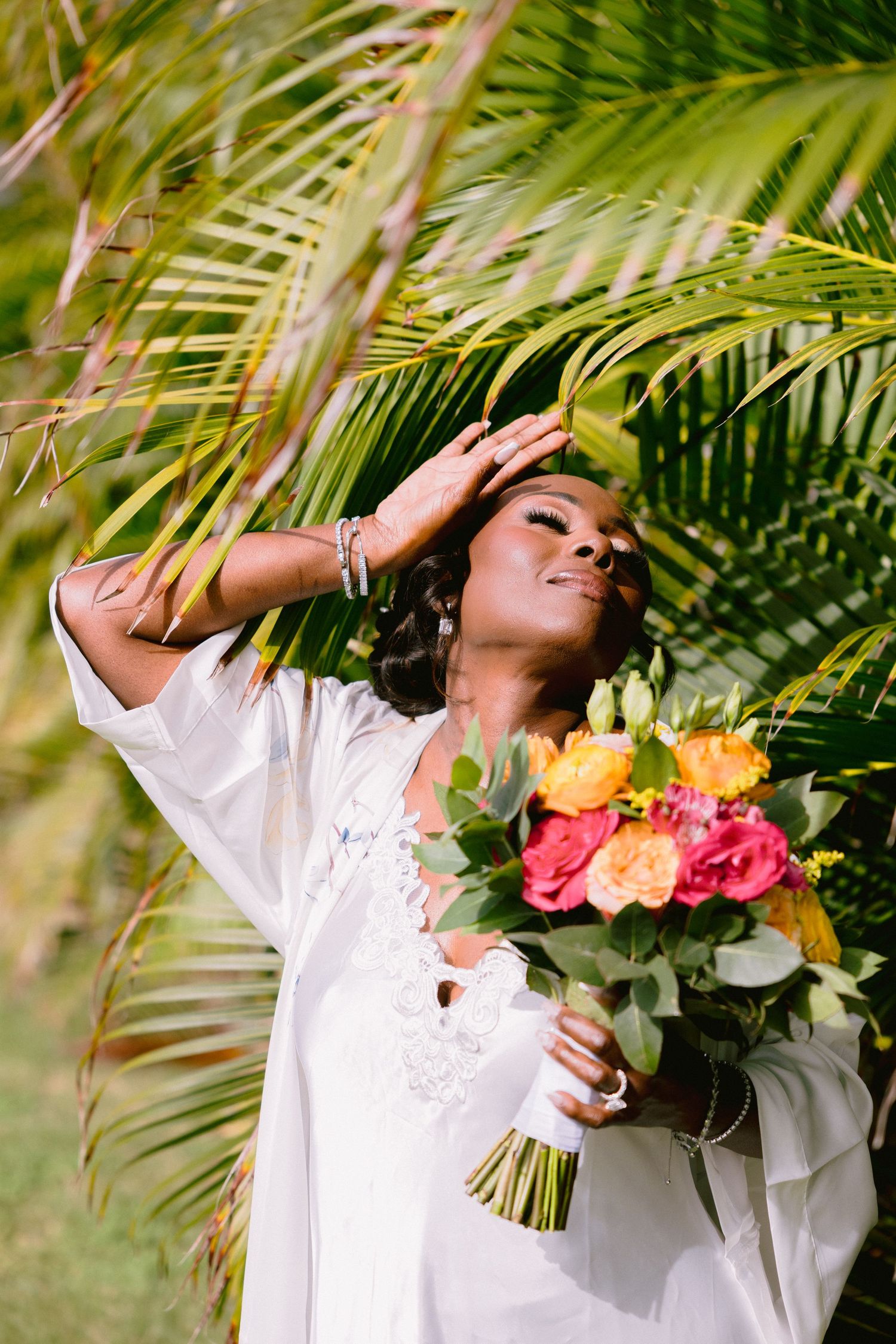 A person in white clothing poses with a colorful bouquet against a backdrop of tropical palm fronds.