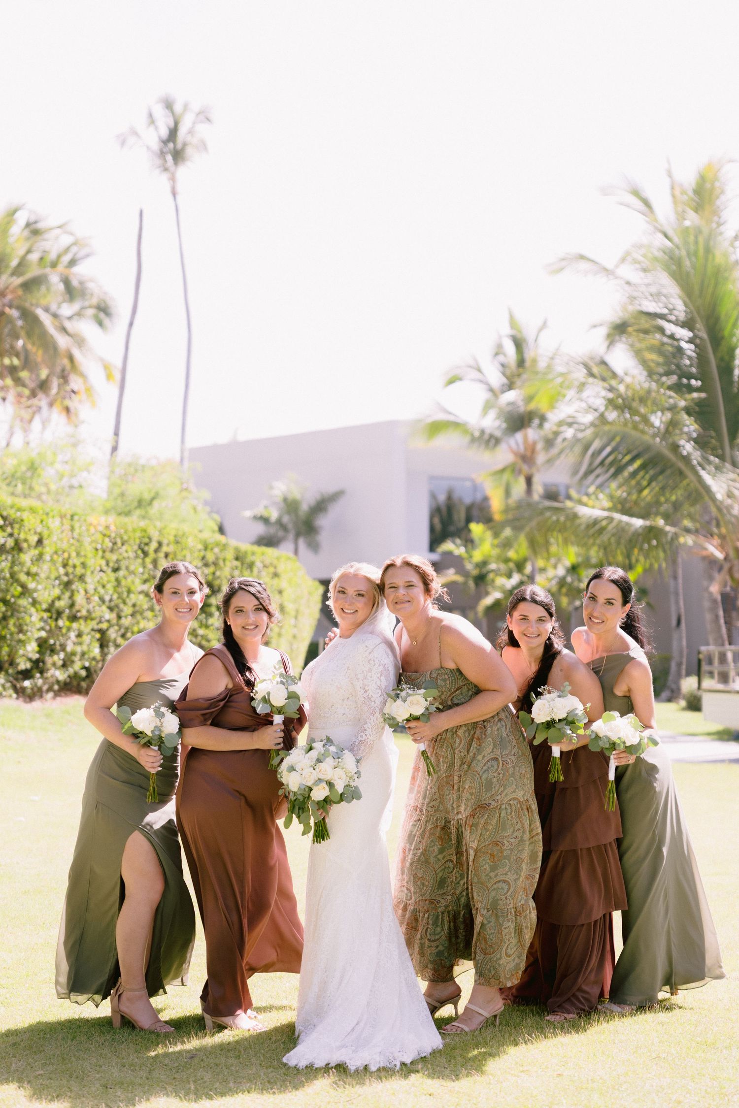 Bride in white lace gown poses with five bridesmaids in earthy tones holding white bouquets at tropical outdoor wedding.