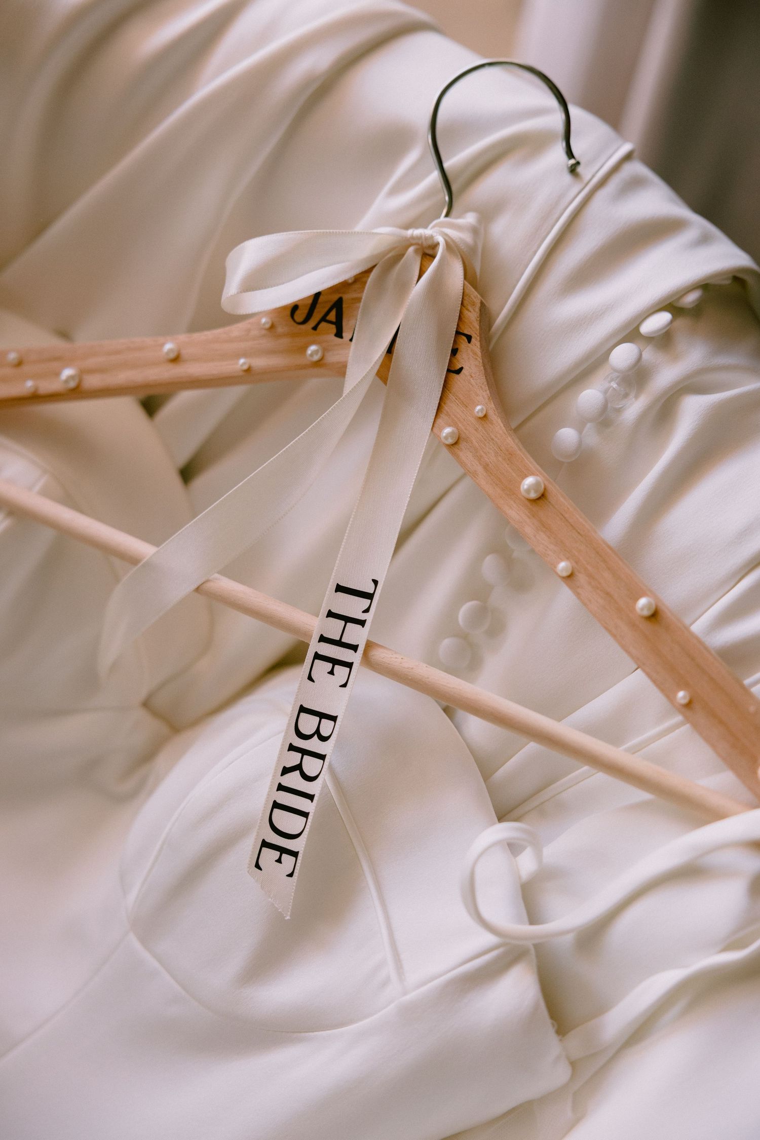 Wooden wedding hangers with 'The Bride' ribbon bow resting on a white pearl-embellished bridal gown.