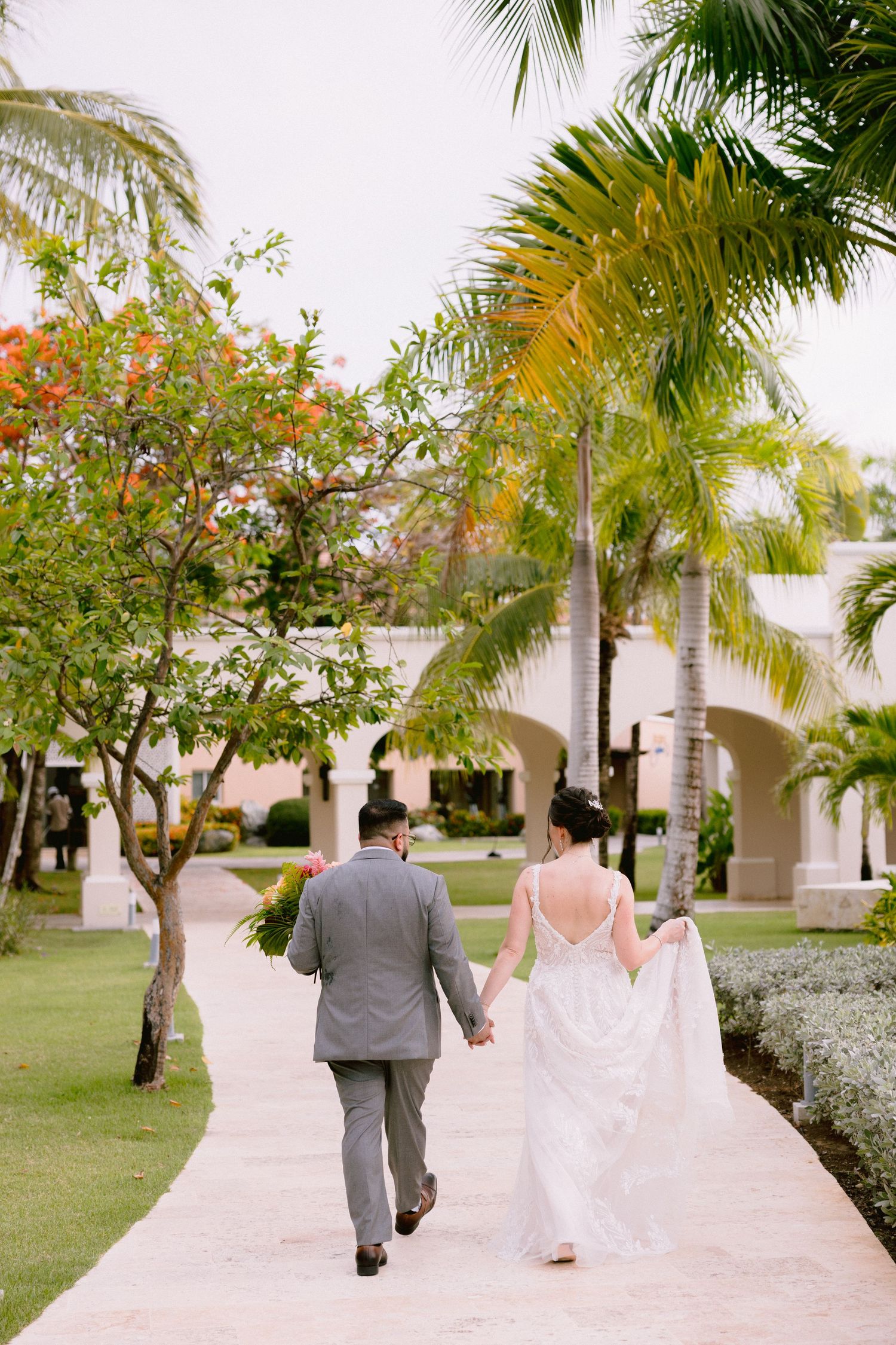 A couple walks hand in hand down a palm-lined path at a tropical resort on their wedding day.