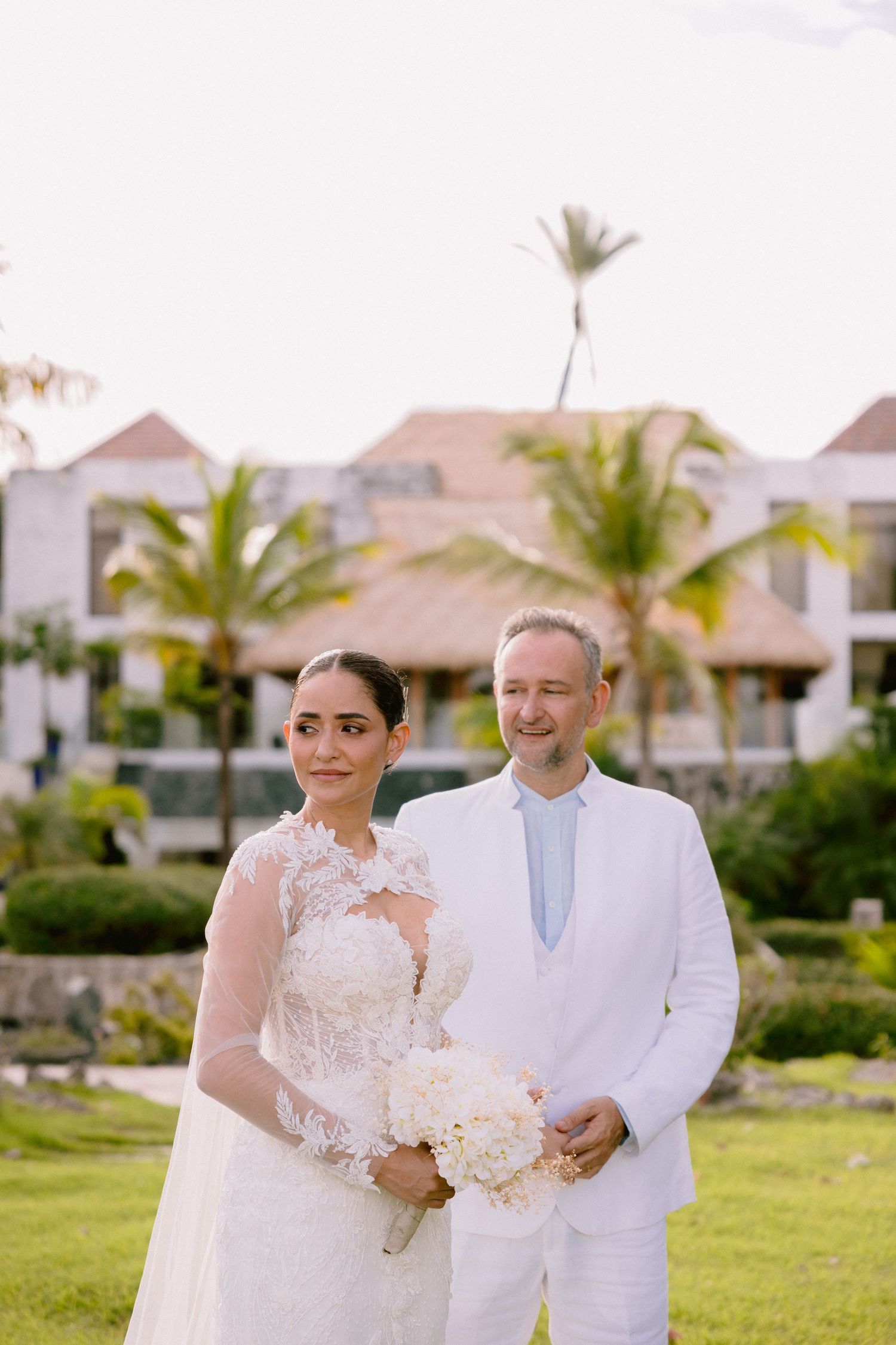A couple in white wedding attire walks together on a grassy lawn with palm trees and buildings in the background.