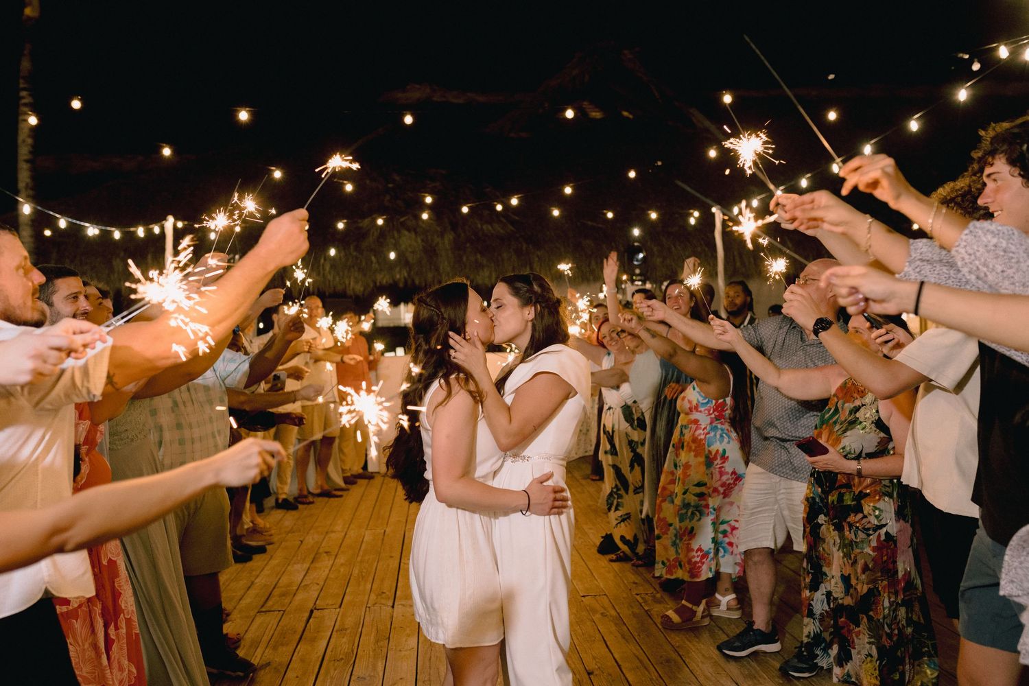 Two brides share a kiss during a romantic sparkler send-off at their outdoor wedding reception at night.