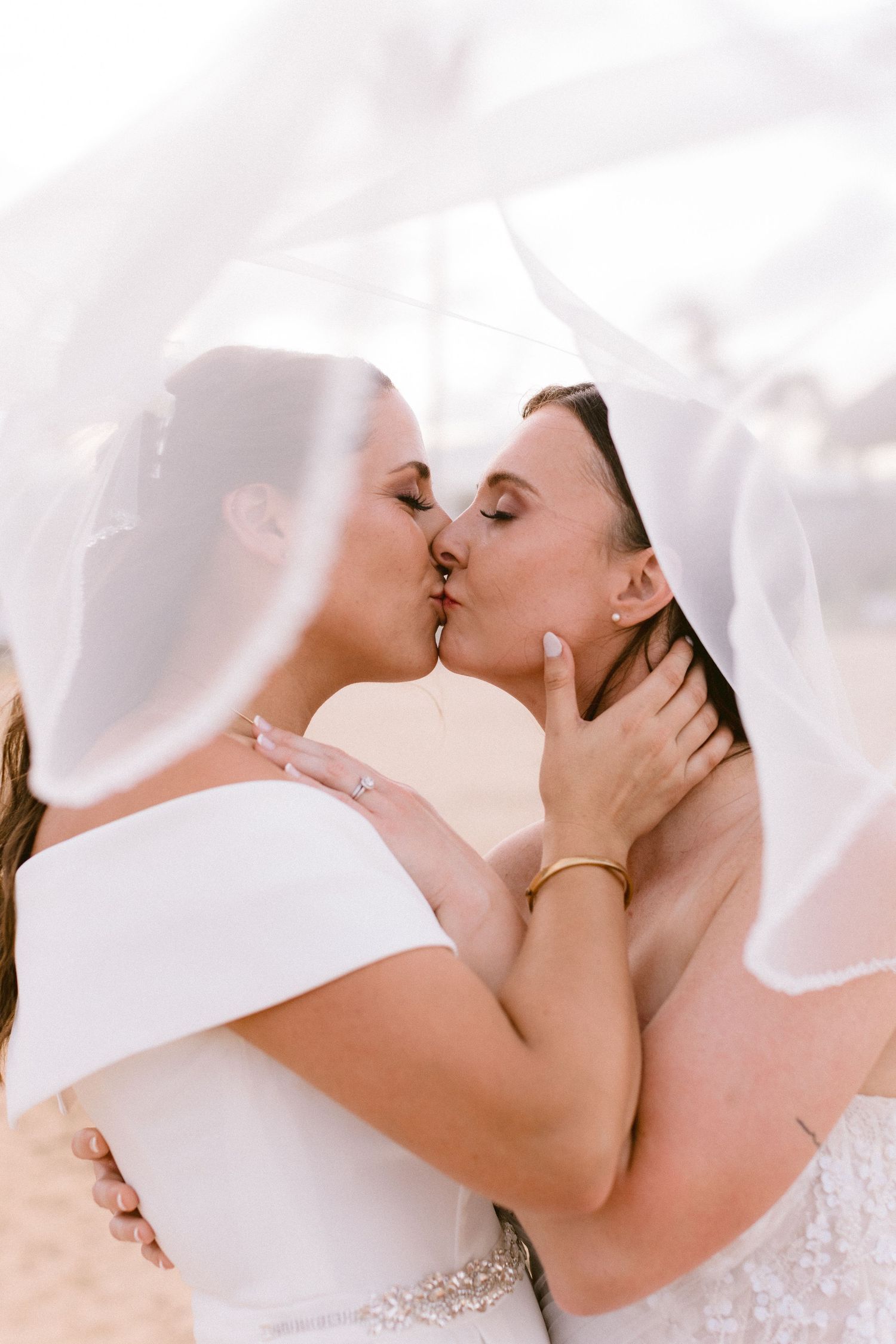 Two brides share a romantic kiss under a flowing white veil on their wedding day.