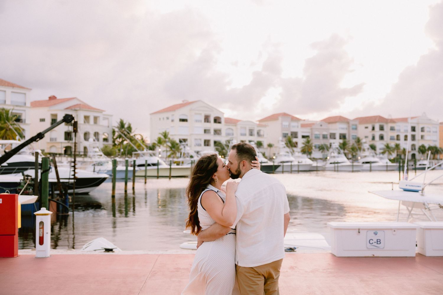 Couple embracing romantically at a marina with white buildings, palm trees, and boats in the background.