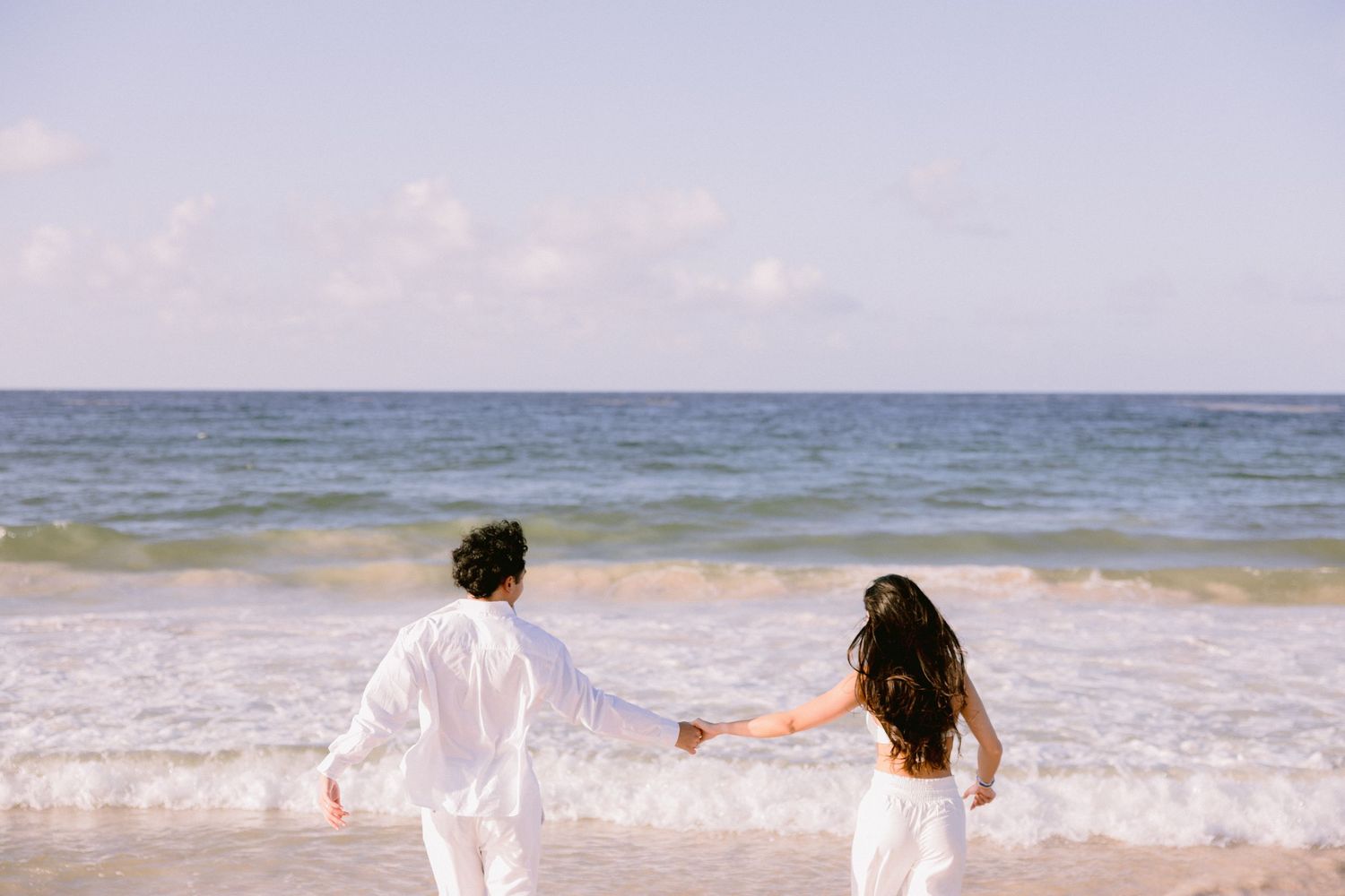 Couple in white holding hands walking toward ocean waves on a sunny beach.