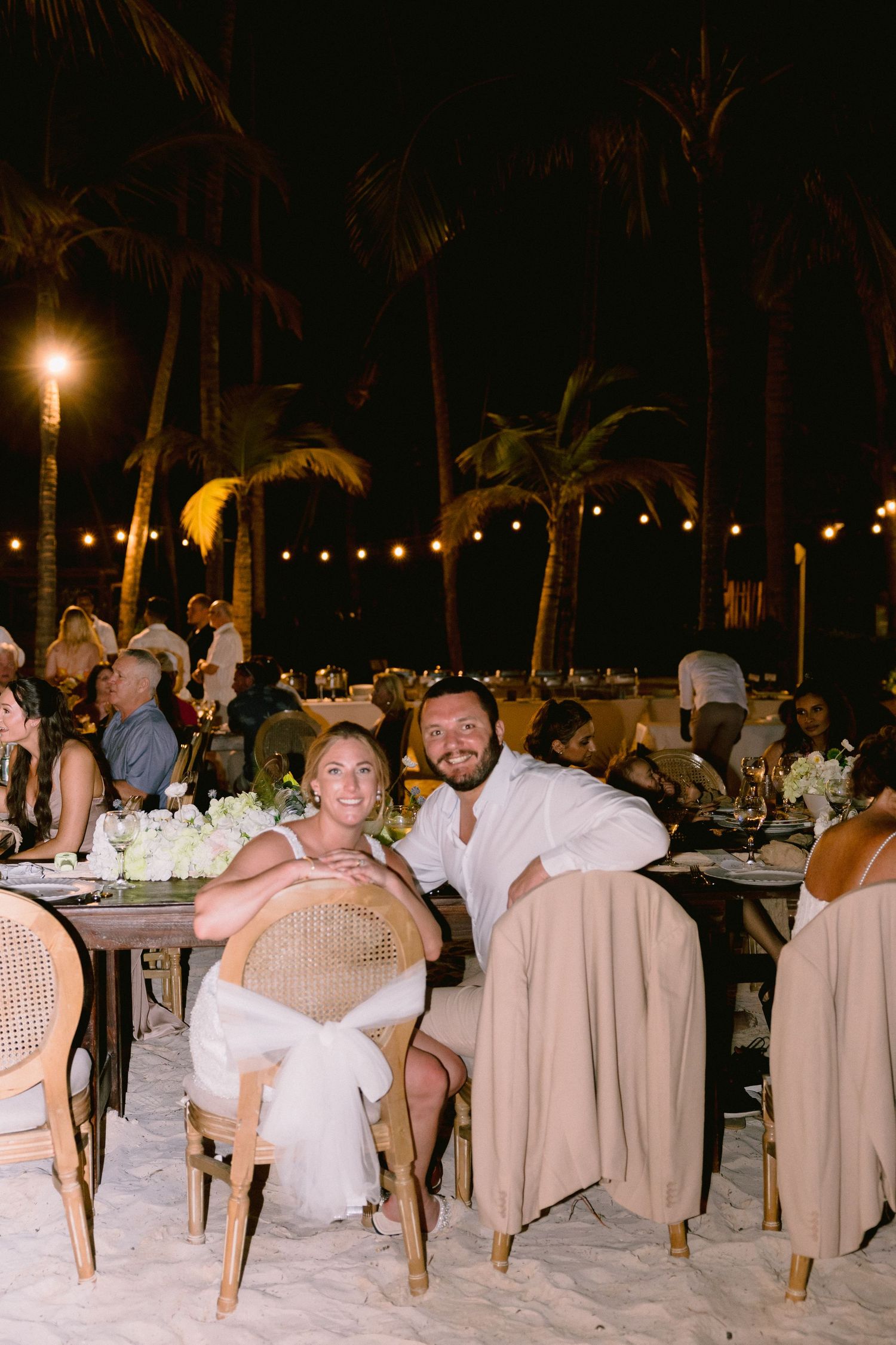 Couple smiling at sweetheart table during outdoor beach wedding reception at night with palm trees and string lights.