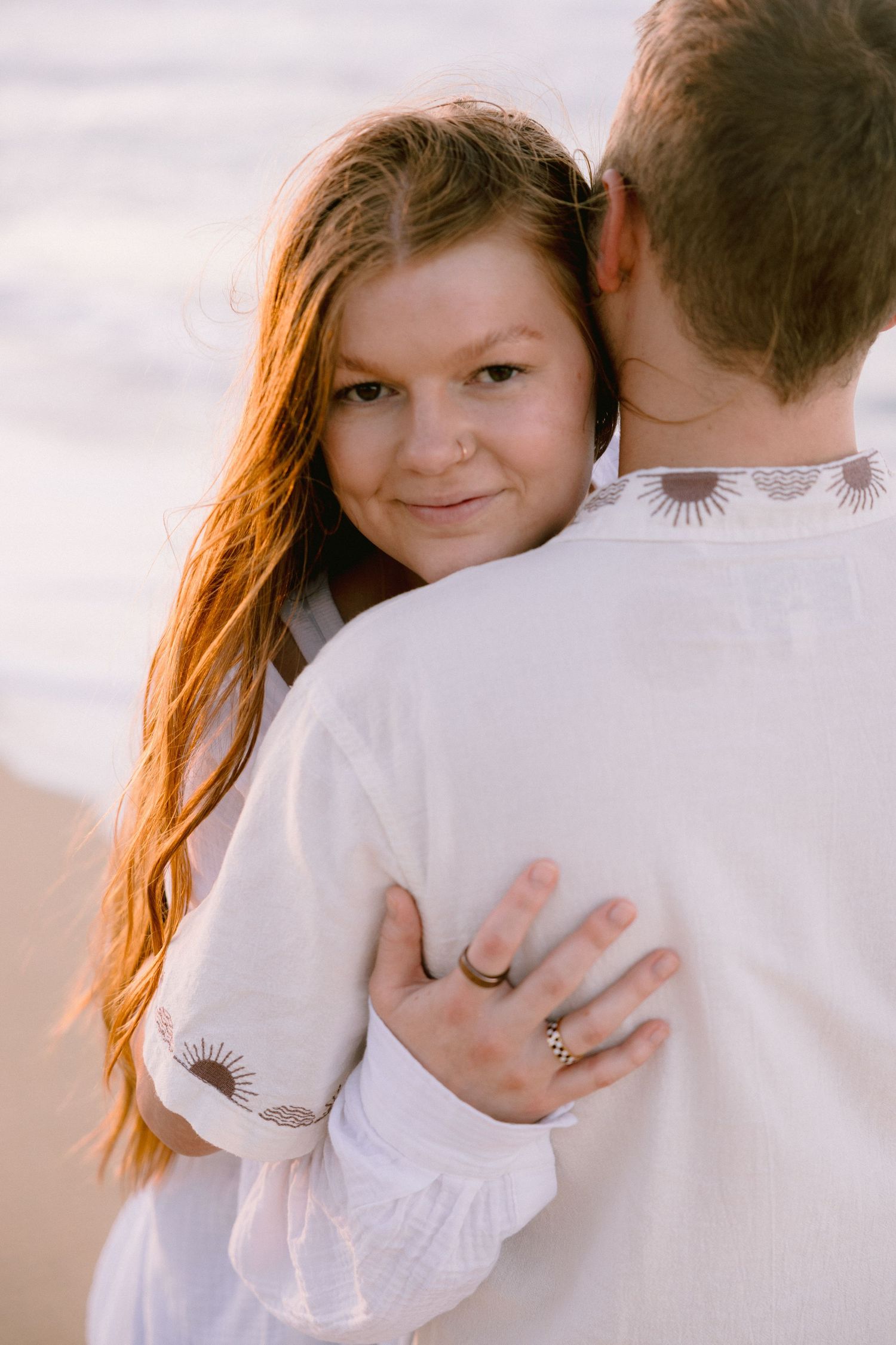 A woman with red hair hugs a man from behind on a beach, smiling warmly while wearing a white dress.