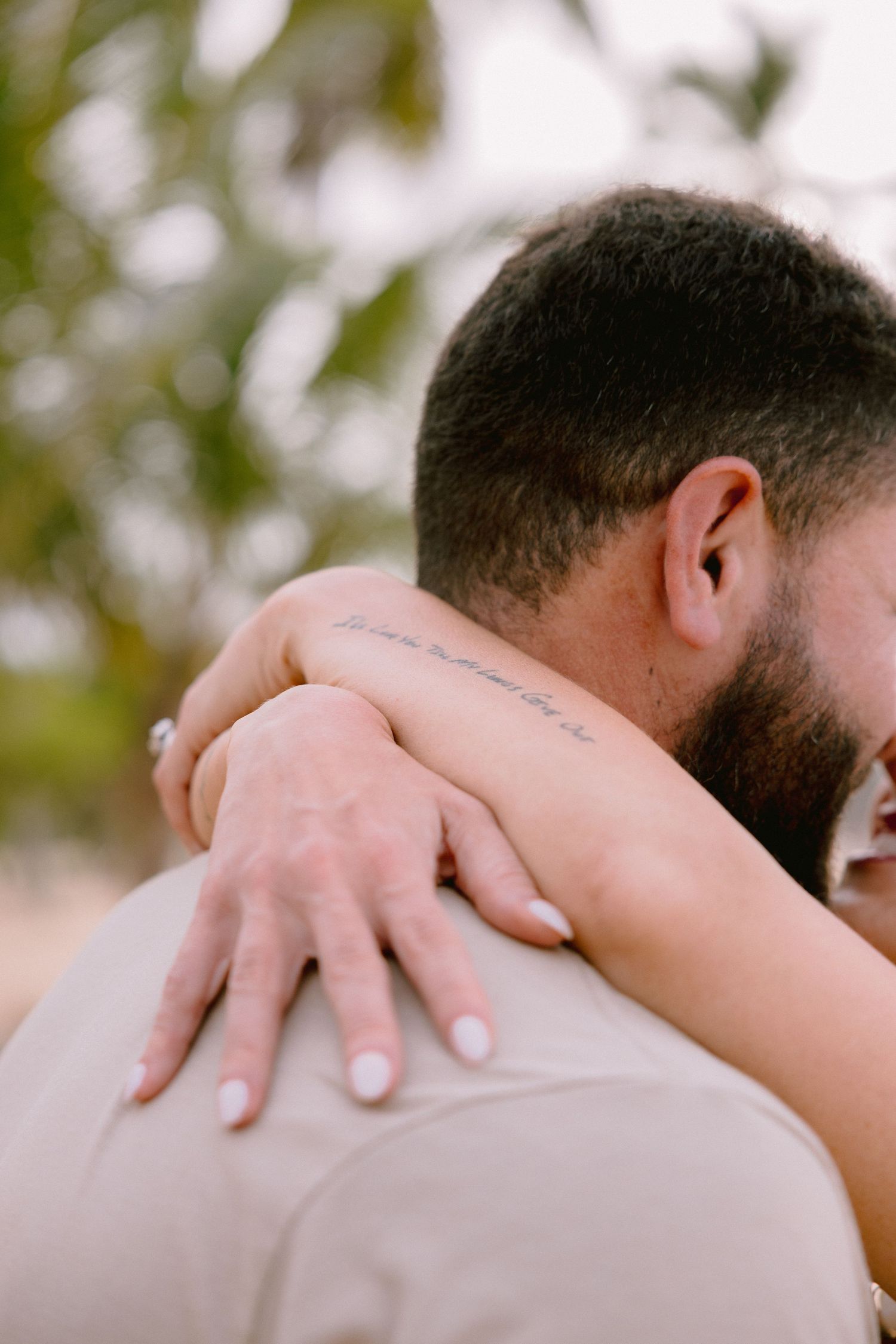 Couple embracing closely, woman's arm with script tattoo wrapped around man's shoulder, engagement ring visible.