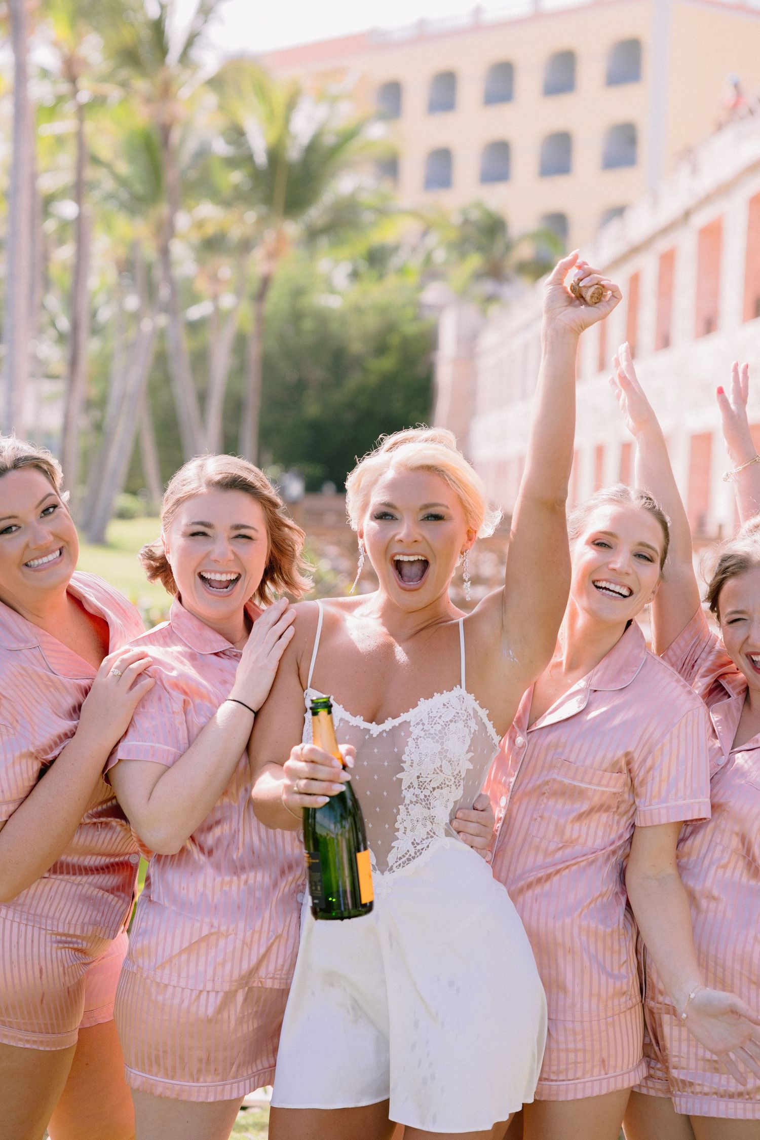 Bride in white lace romper celebrates with bridesmaids in pink outfits, holding champagne bottle outdoors near palm trees.