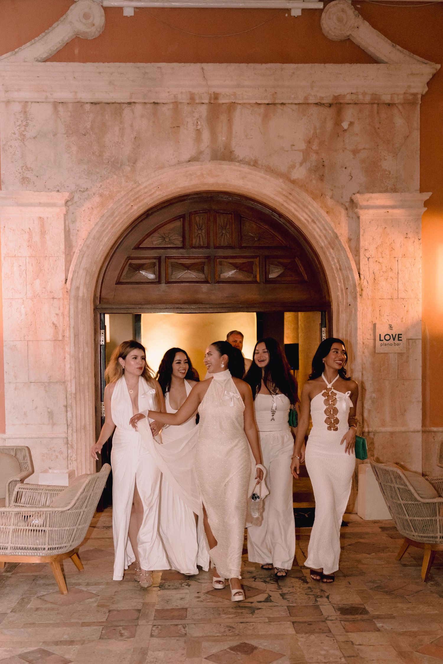 Five women in elegant white dresses walk arm in arm through a grand stone archway at an evening wedding celebration.