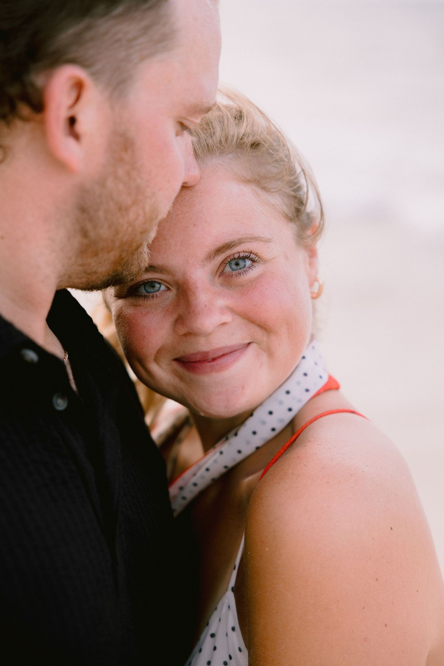 A smiling blue-eyed woman with freckles leans close to a man in a black shirt in a romantic outdoor portrait.