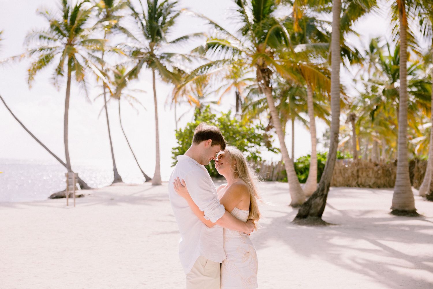 A couple embraces on a pristine white sand beach lined with tall palm trees in tropical paradise.