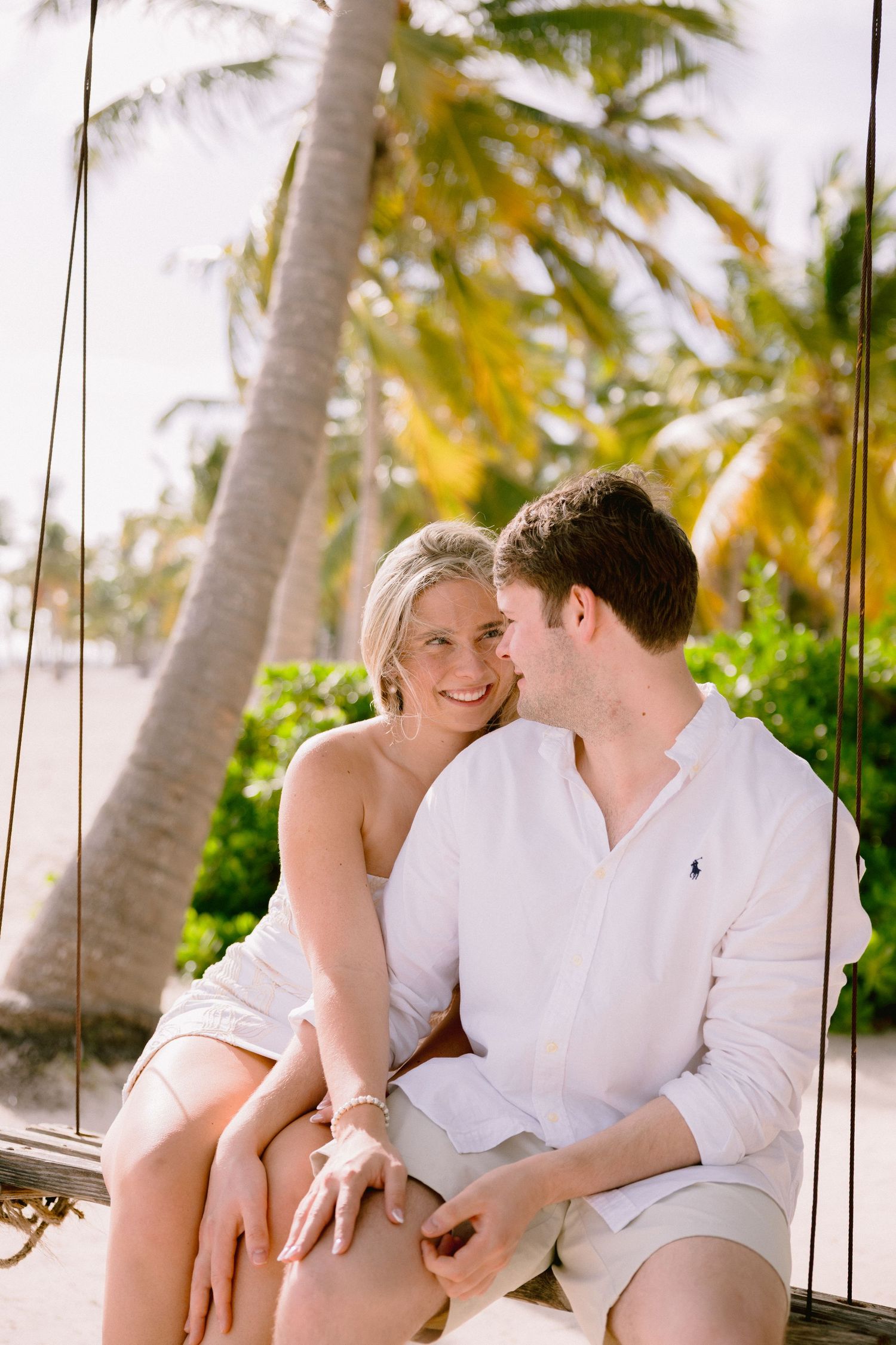 A romantic couple in white clothing share tender moments on a tropical beach with palm trees in the background.