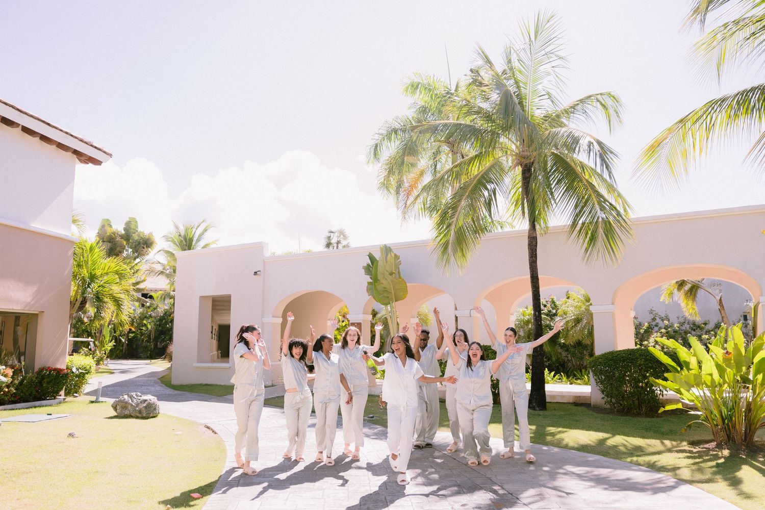 Wedding party in white attire celebrating outdoors at a tropical resort with palm trees and pink archways.