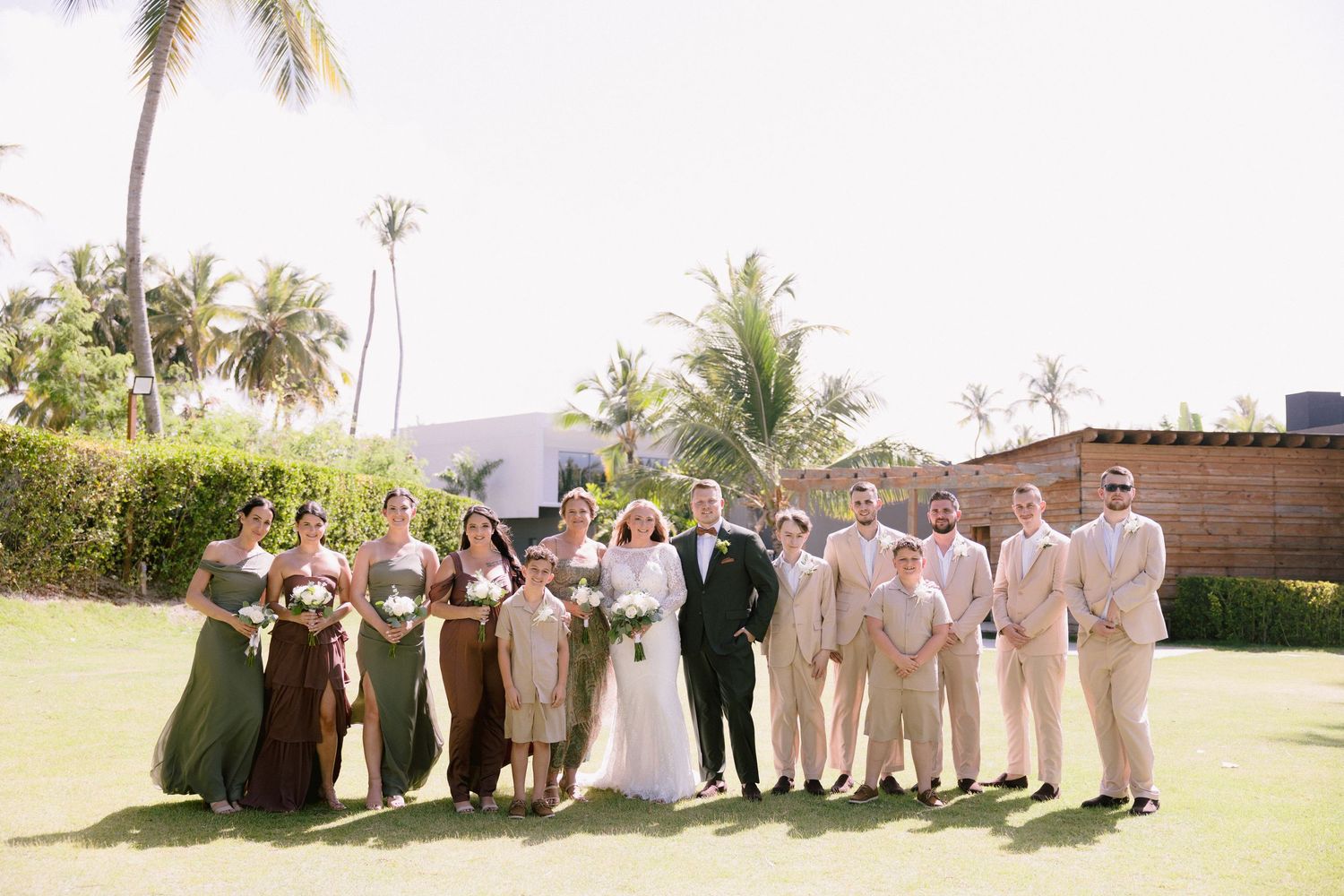 Wedding party posing outdoors on green lawn with palm trees, bridesmaids in green dresses and groomsmen in tan suits.