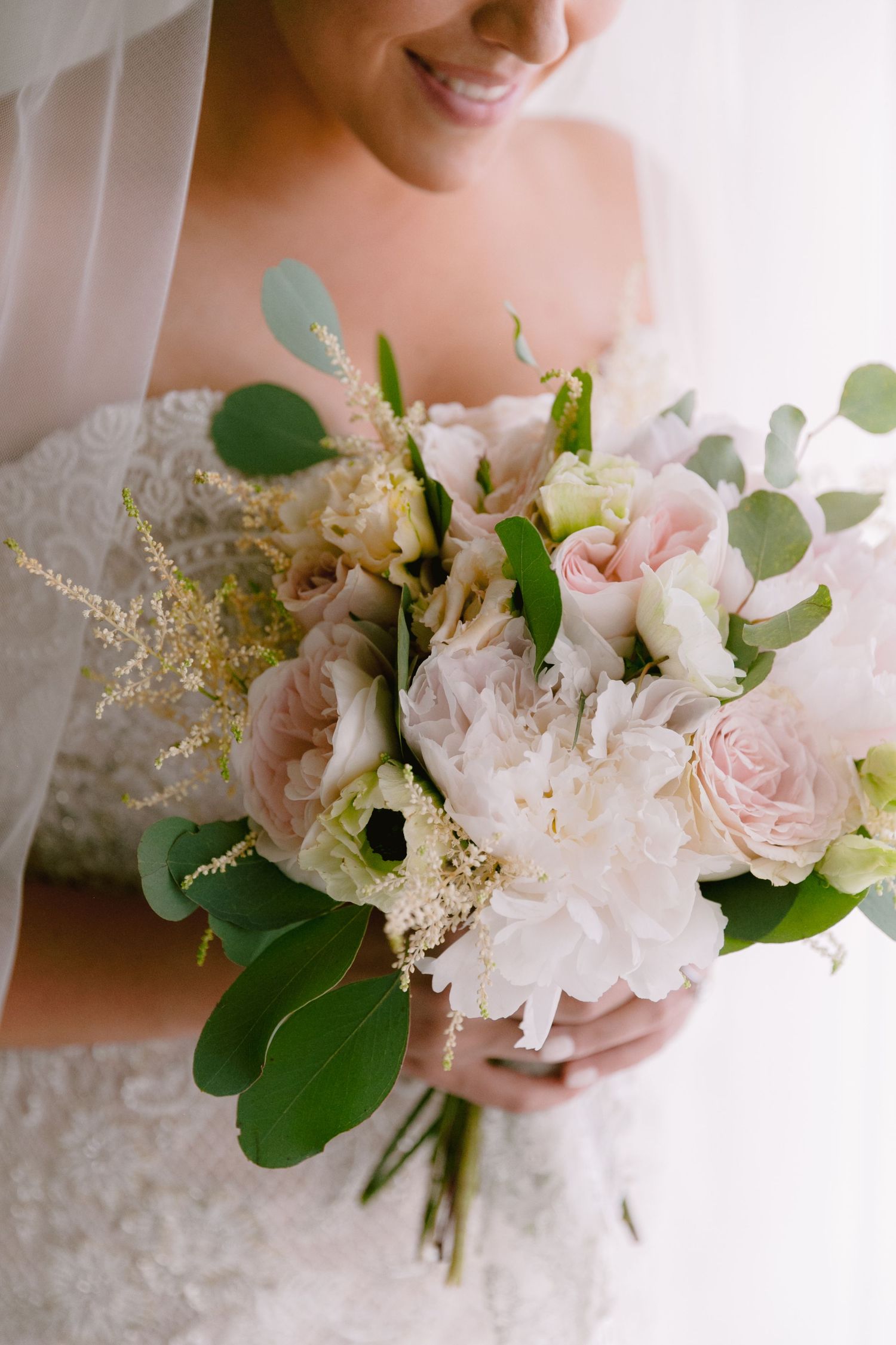Romantic bridal bouquet featuring blush roses, white peonies, and eucalyptus leaves held against a lace wedding dress.