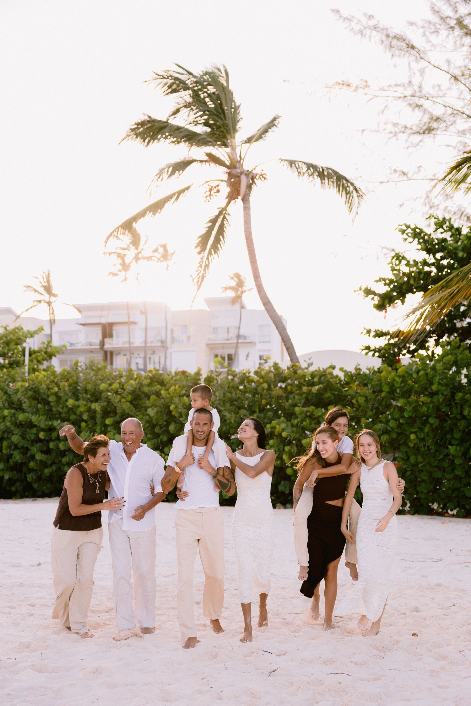 A group plays and laughs together on a beach with palm trees swaying in the background.