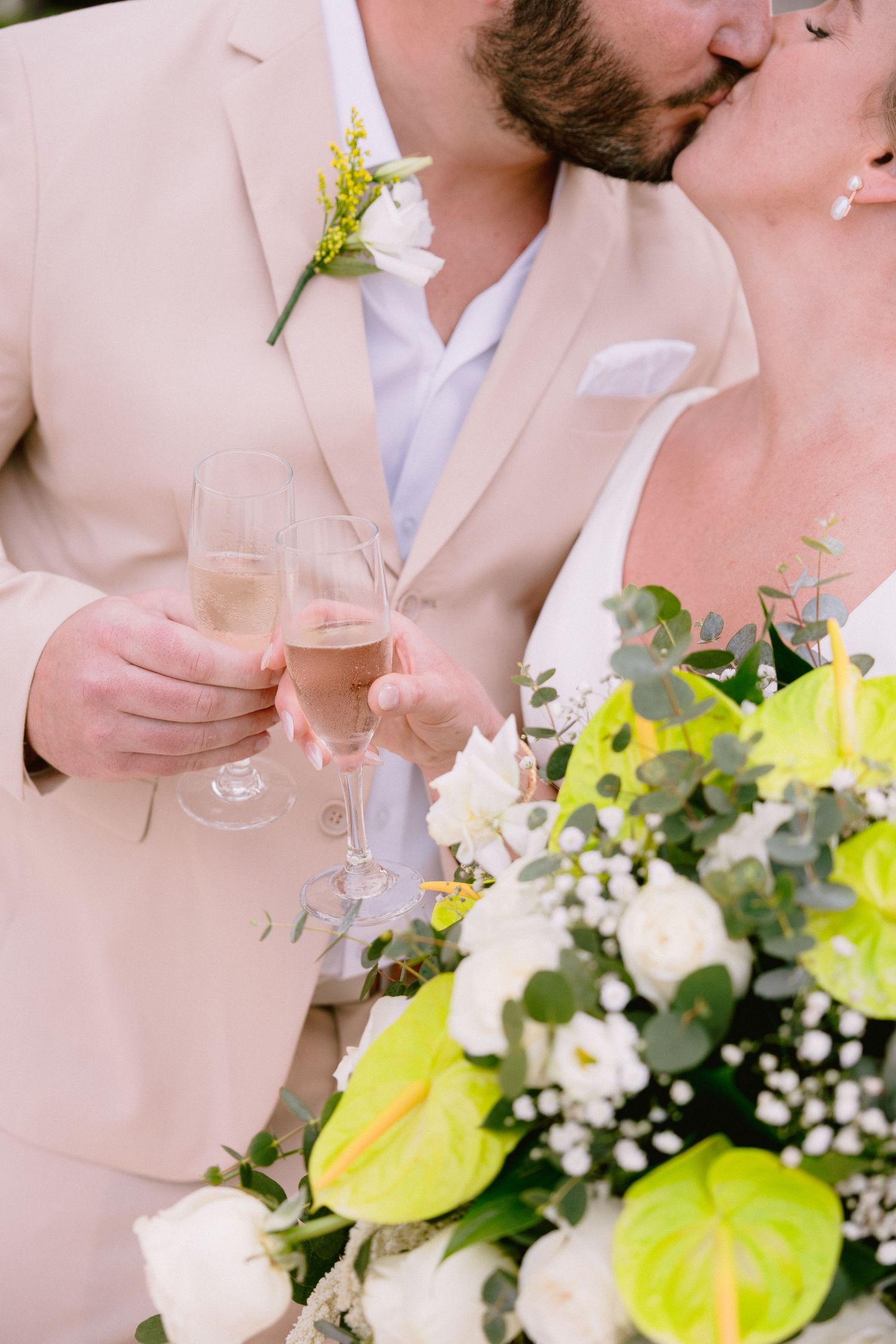Newlyweds kissing while toasting champagne glasses, bride holding bouquet of white roses and green anthuriums.