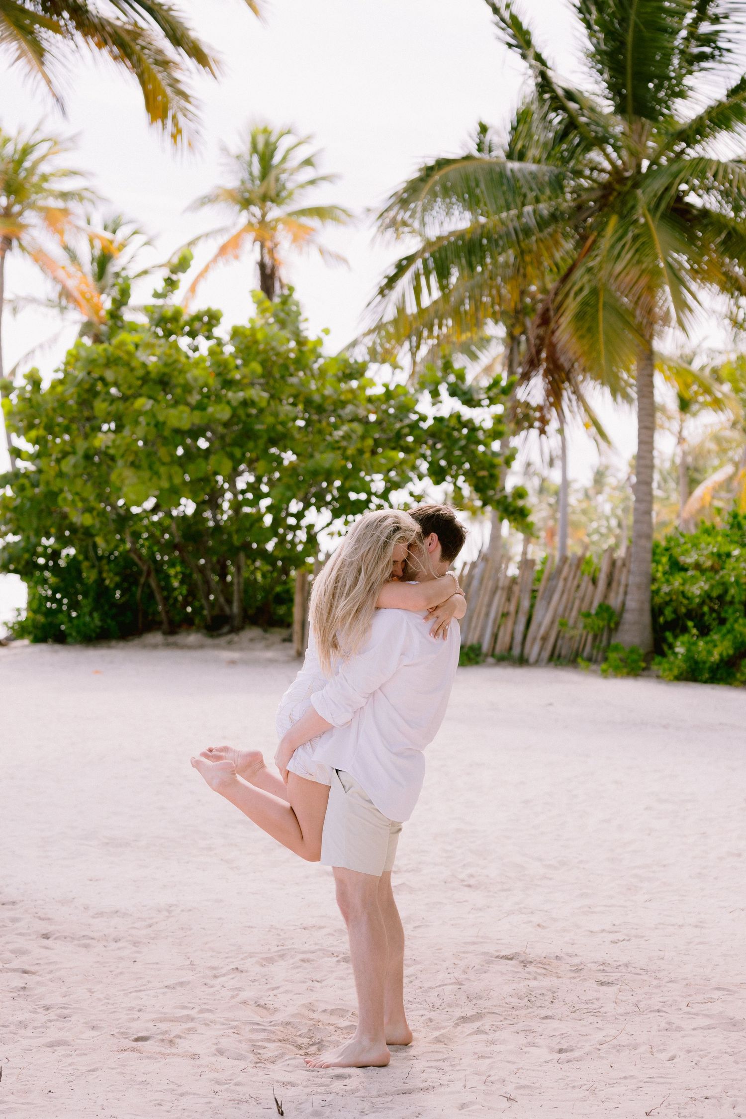 A couple embraces on a tropical beach with palm trees and white sand creating a romantic paradise setting.