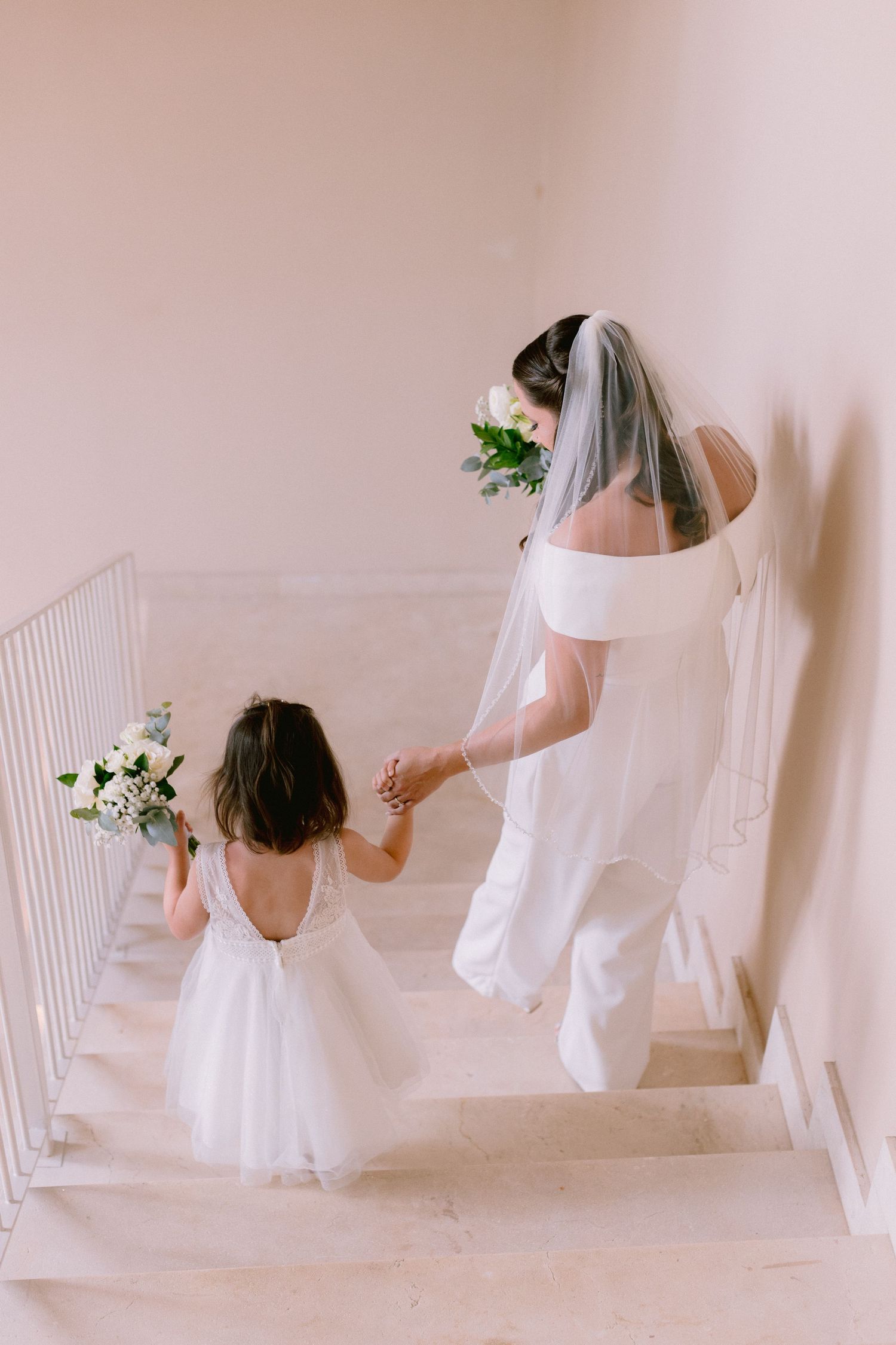 Bride in white gown and veil holds hands with flower girl in white dress while walking up stairs holding bouquets.