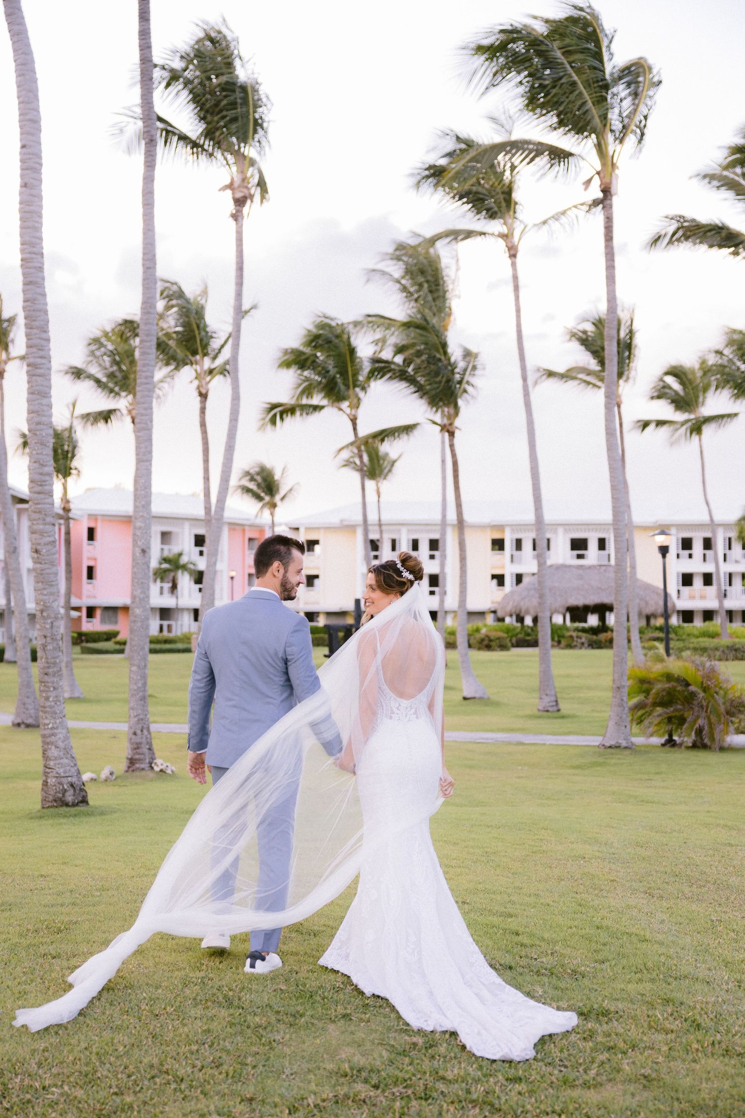 A couple in wedding attire walks on a palm-lined lawn with white buildings in the background on a breezy day.