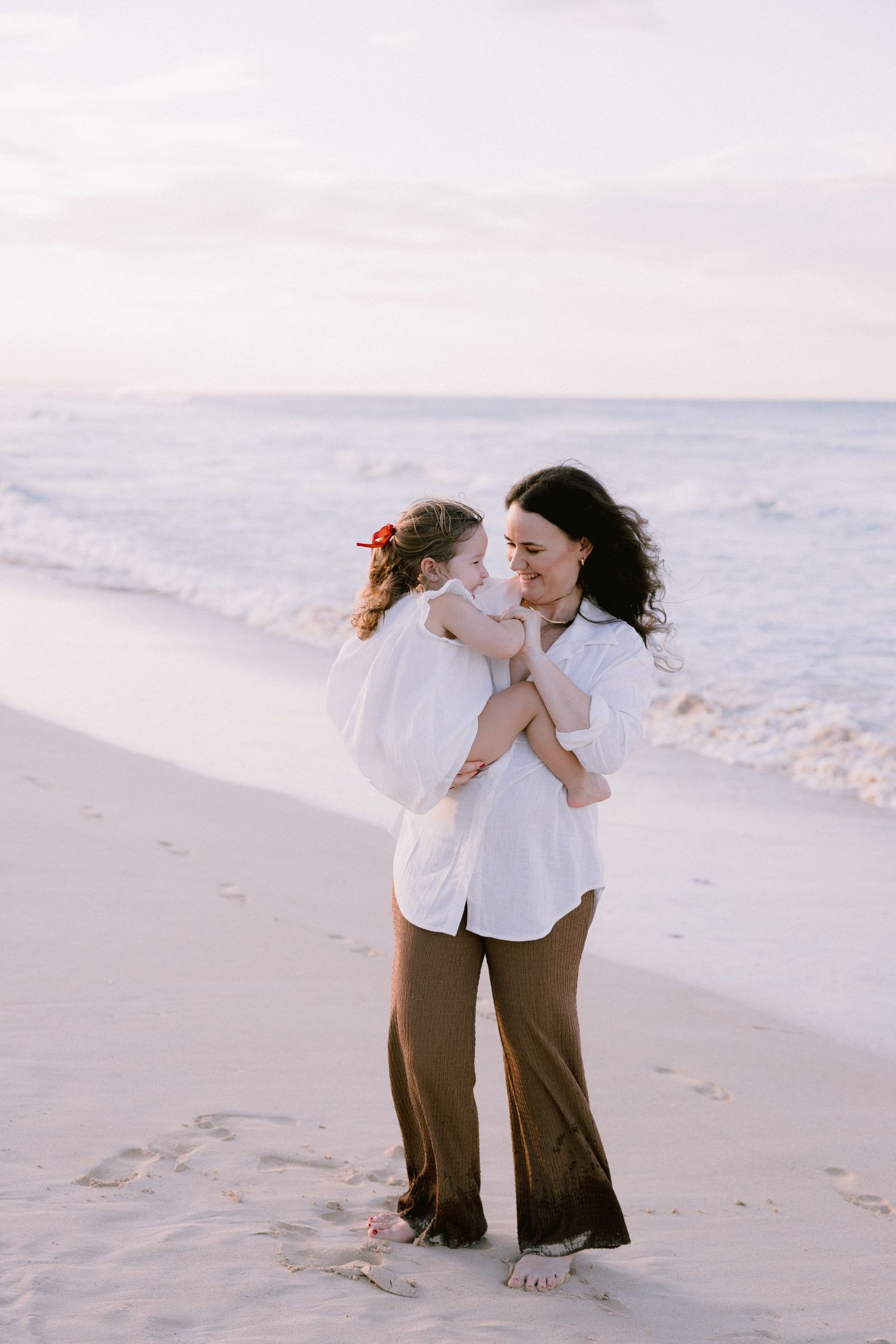 Two people in white and brown outfits share tender moments while walking along a serene beach at sunset.