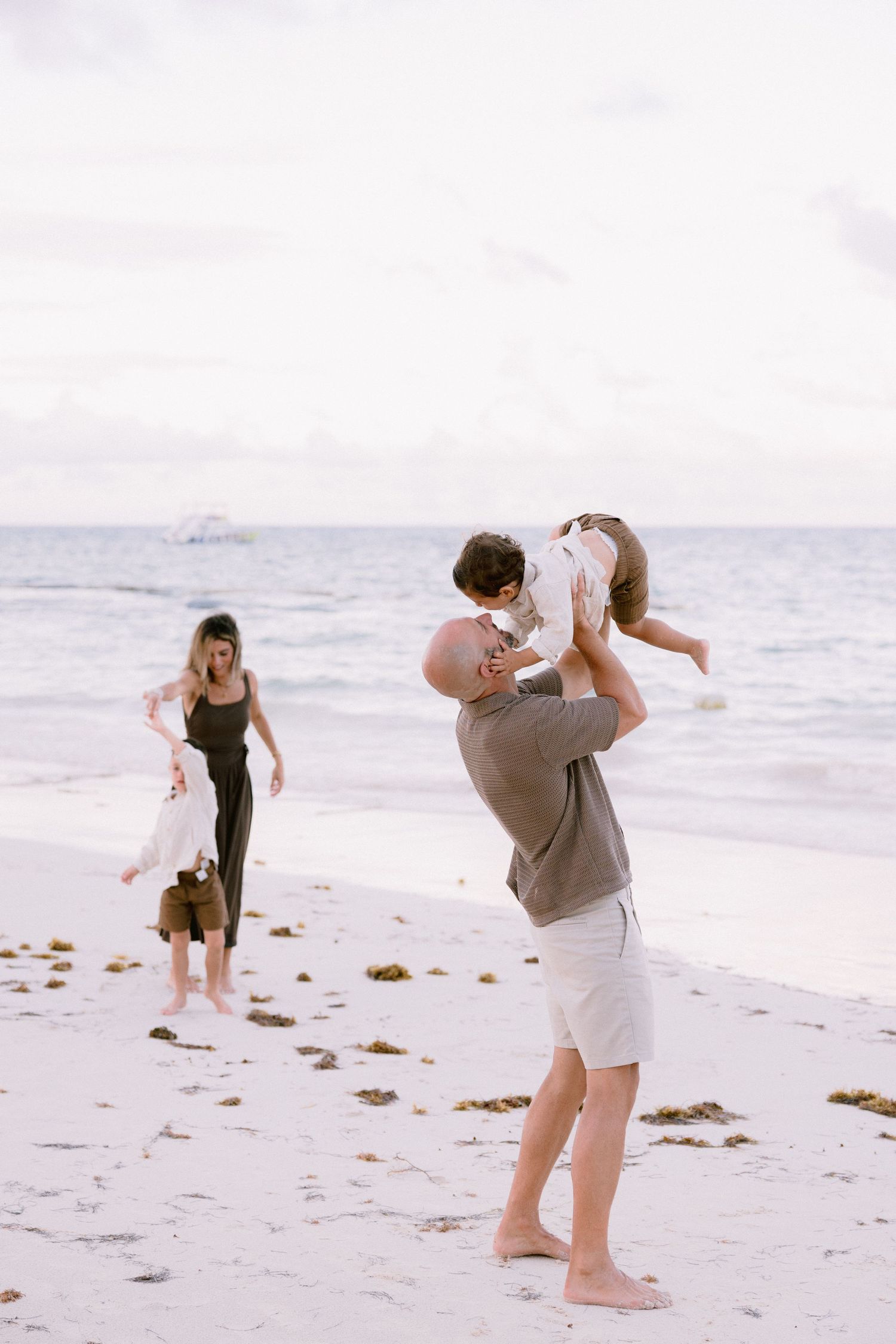 Family enjoying a beach day, with a man lifting a baby in the air while a woman walks nearby on white sand.