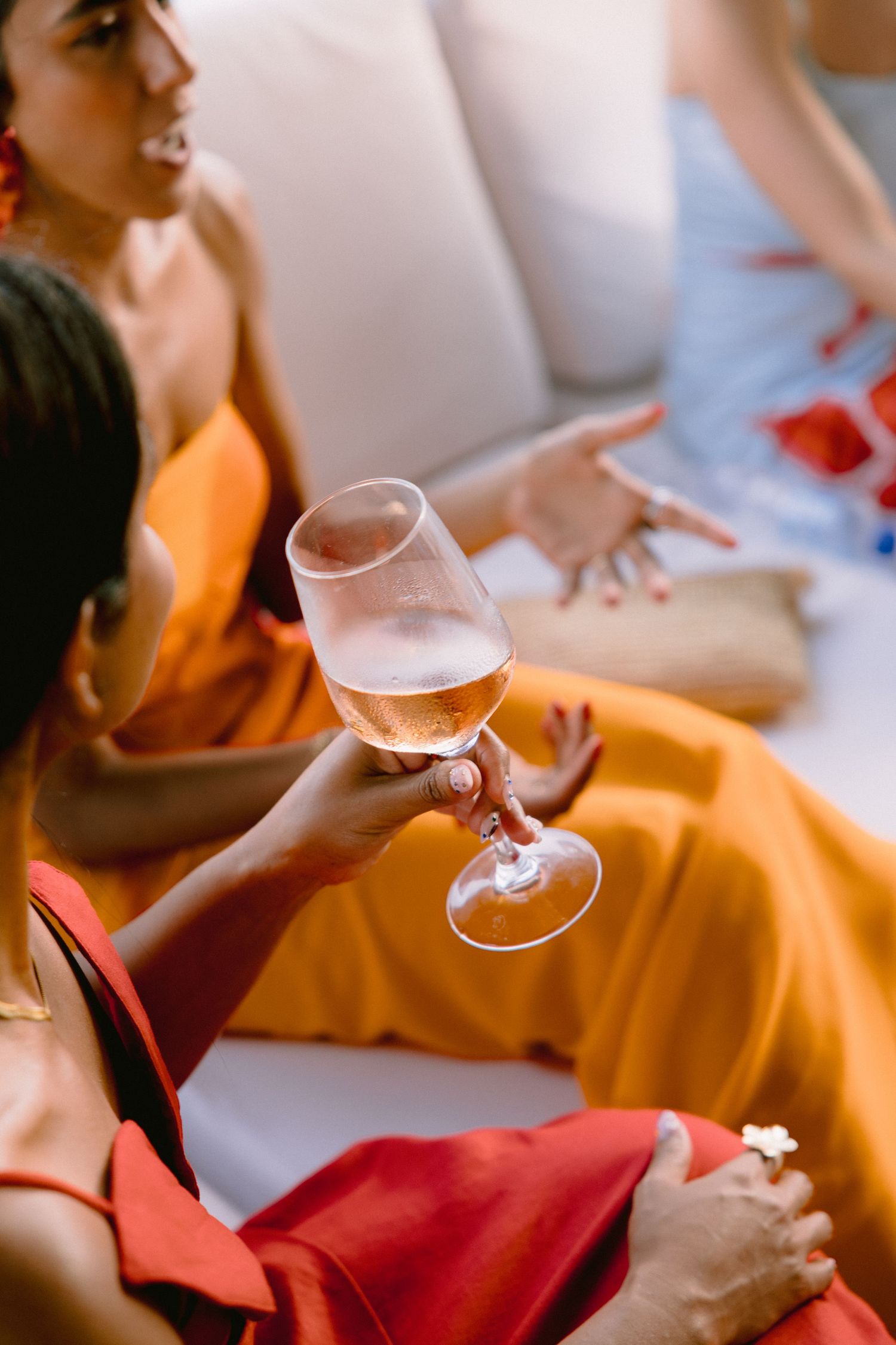 Woman in a red dress holds a glass of rosé wine while a friend in an orange dress gestures on a cream sofa.