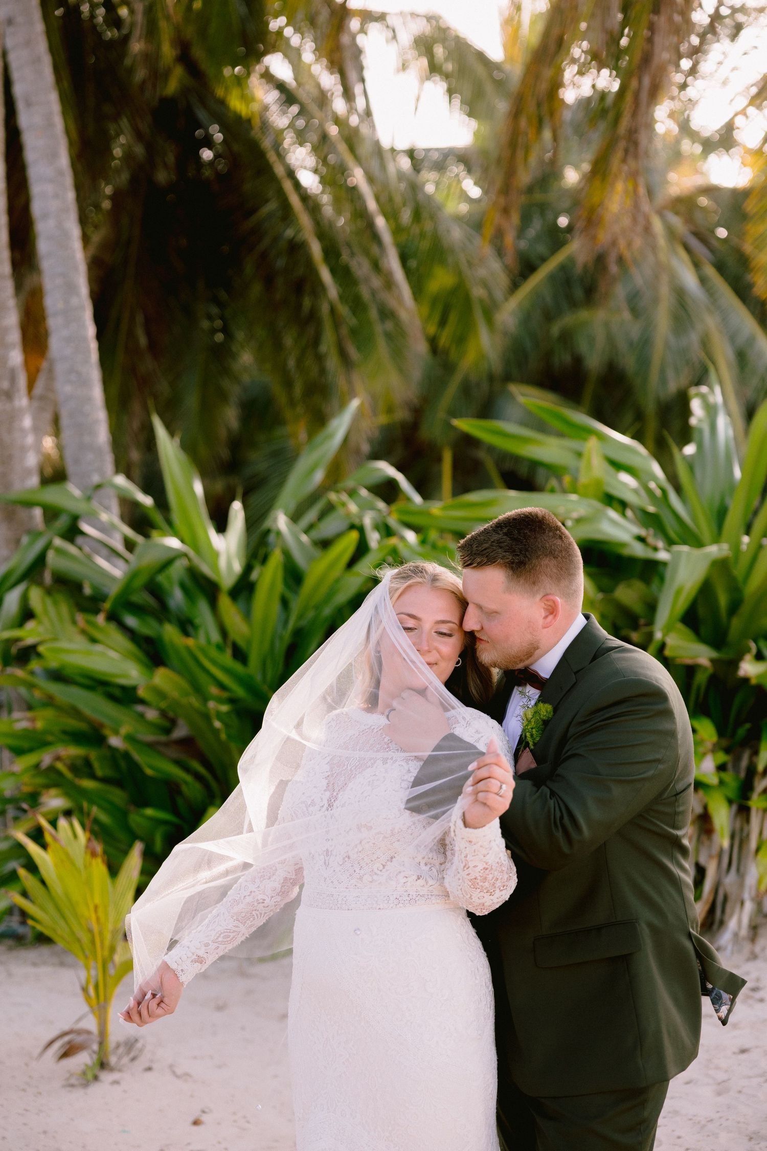 Bride and groom share a romantic moment on a tropical beach, surrounded by lush palm trees in wedding attire.