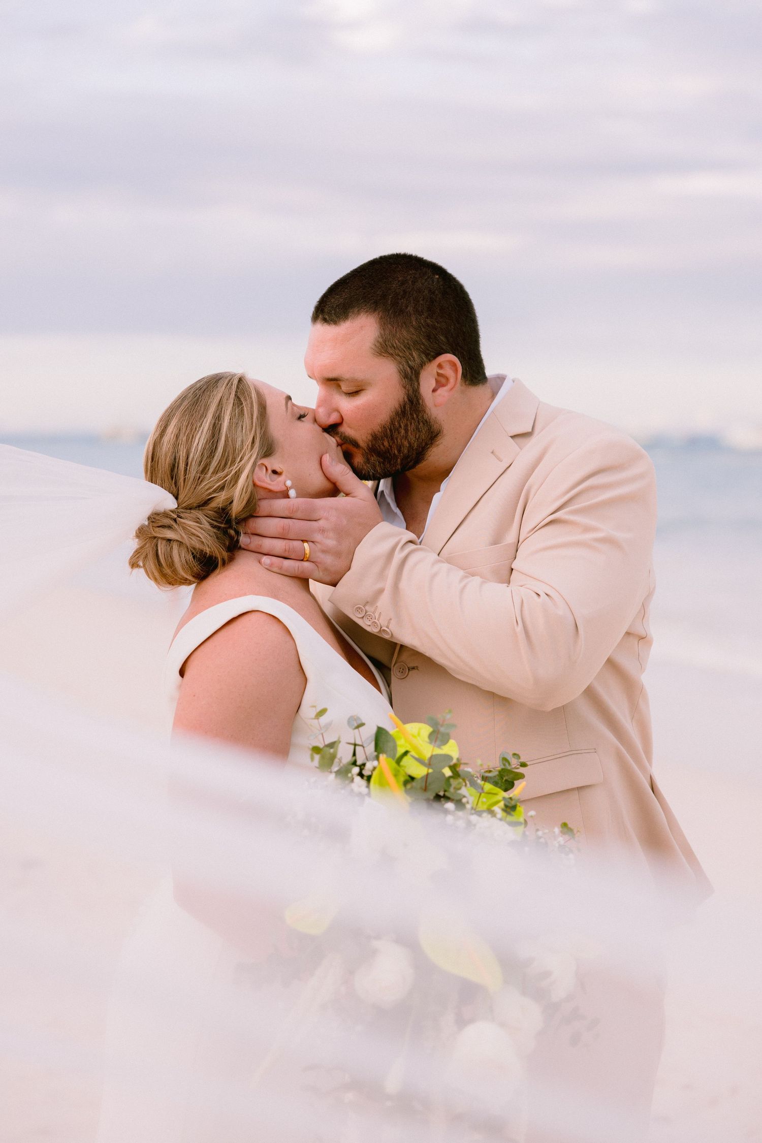 Bride and groom share a romantic first kiss outdoors, with the bride holding a yellow and white bouquet.