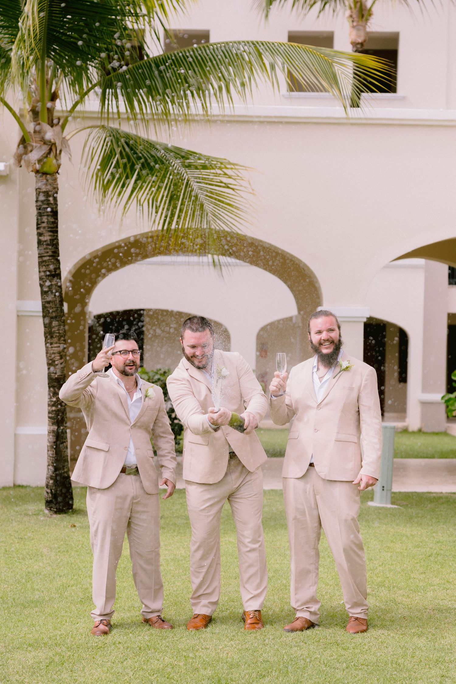 Three groomsmen in tan suits celebrate outdoors by popping champagne near a tropical palm tree and arched building.