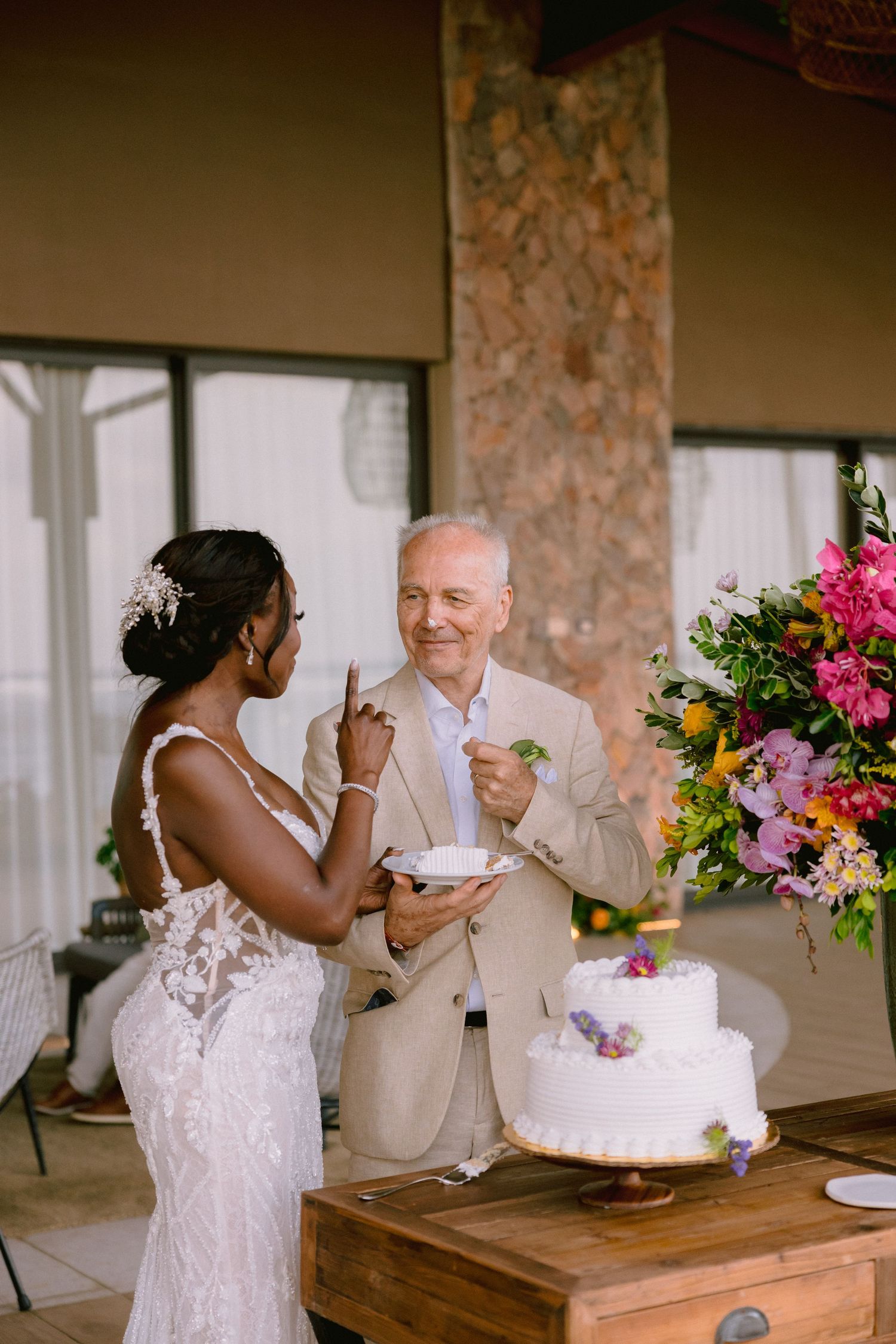 A couple shares cake during a wedding reception next to a colorful floral arrangement on a wooden table.