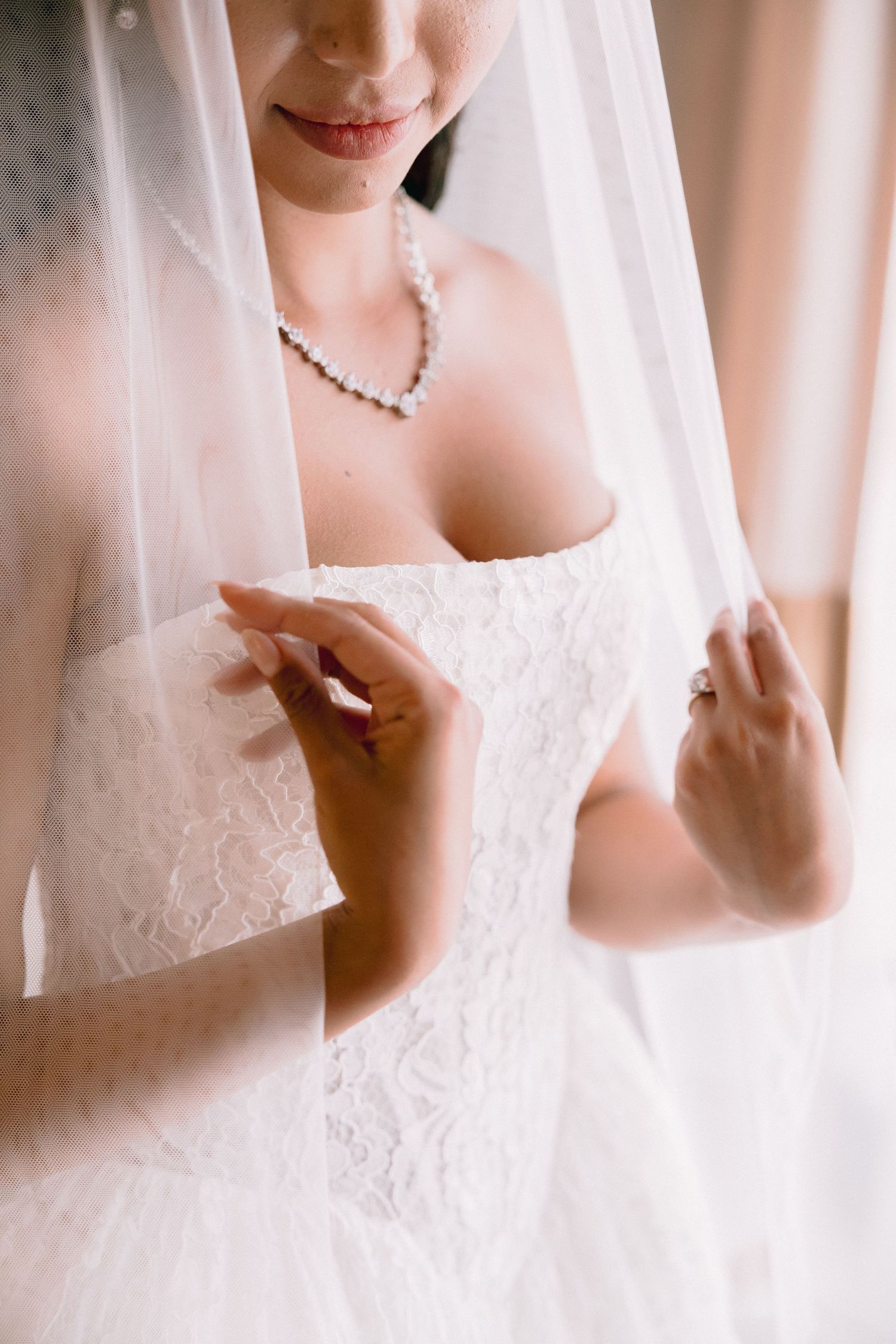 Bride in white lace wedding dress and veil adjusting her gown, wearing pearl necklace and diamond ring.