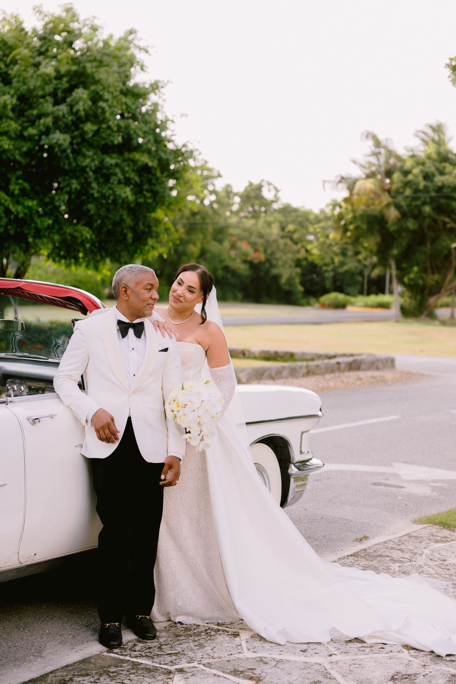 A couple in wedding attire poses elegantly beside a vintage white car on a tree-lined road.