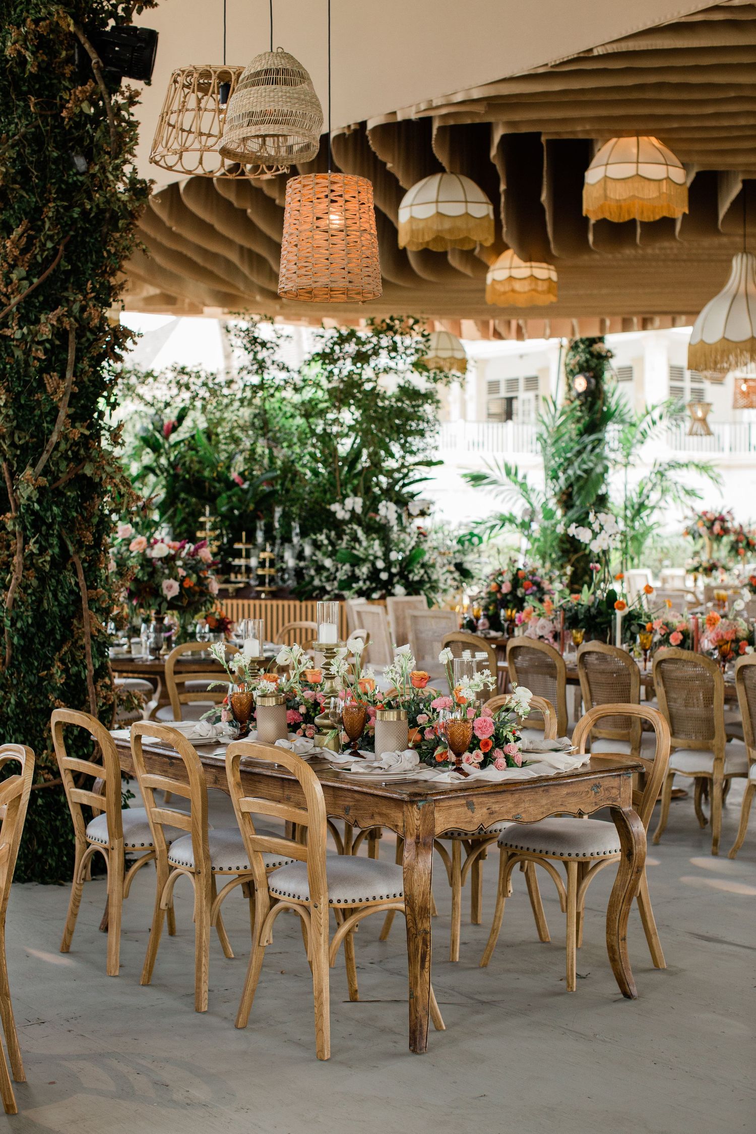 Rustic wedding reception venue with wooden chairs and tables decorated with greenery and floral arrangements under string lights.
