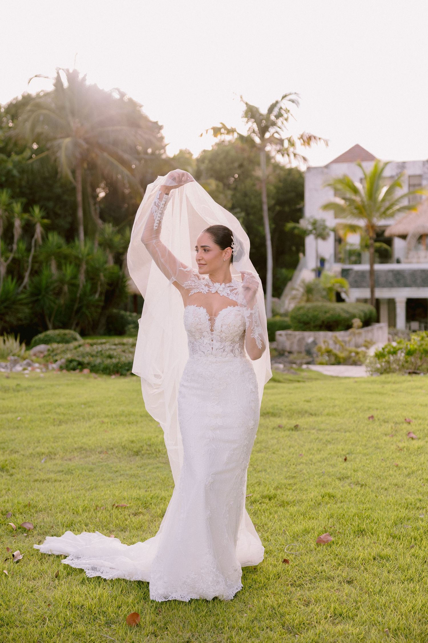 A bride in a flowing white lace wedding dress twirls gracefully on a sunlit green lawn during golden hour.