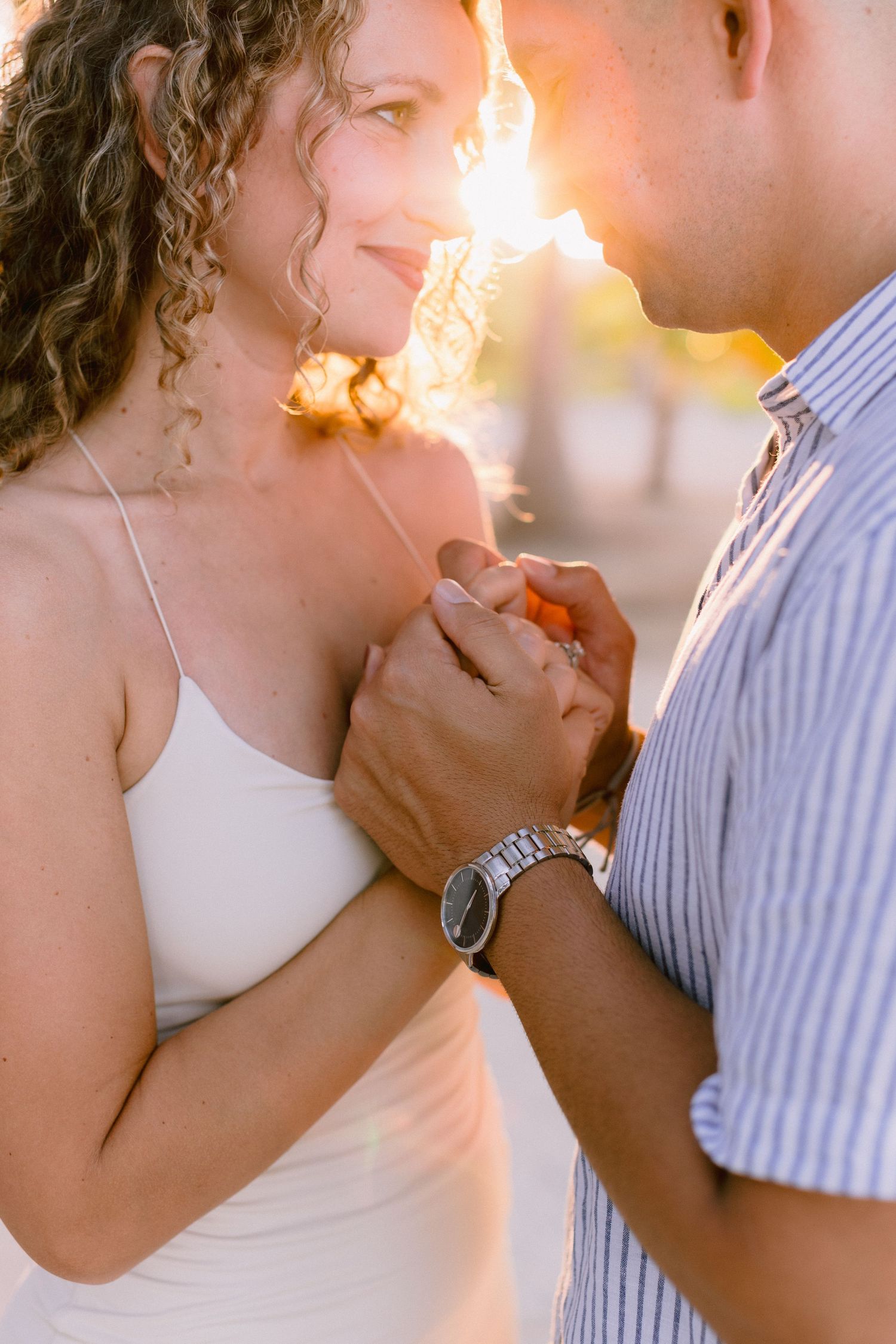 A couple holds hands tenderly at golden hour, the woman in a white dress smiling as warm sunlight glows behind them.