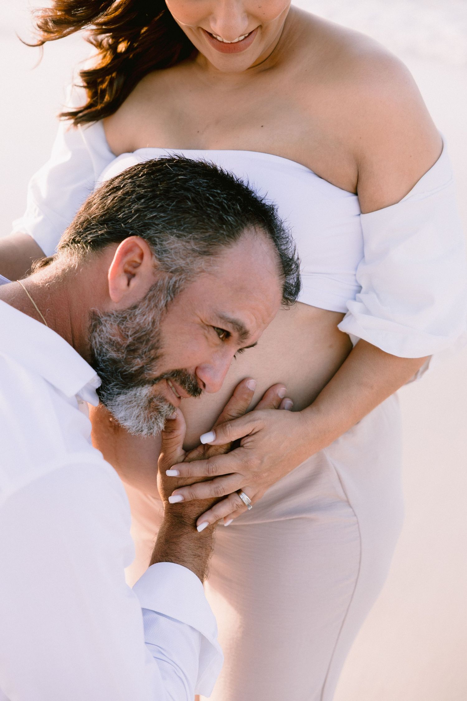 A man with a gray beard tenderly kisses a pregnant woman's belly outdoors, both dressed in white.