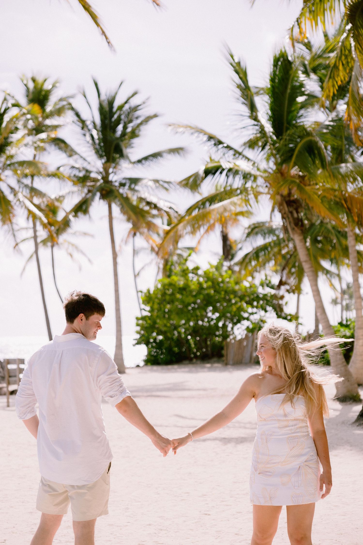 Couple holding hands on a tropical beach with palm trees, both dressed in white outfits on white sand.