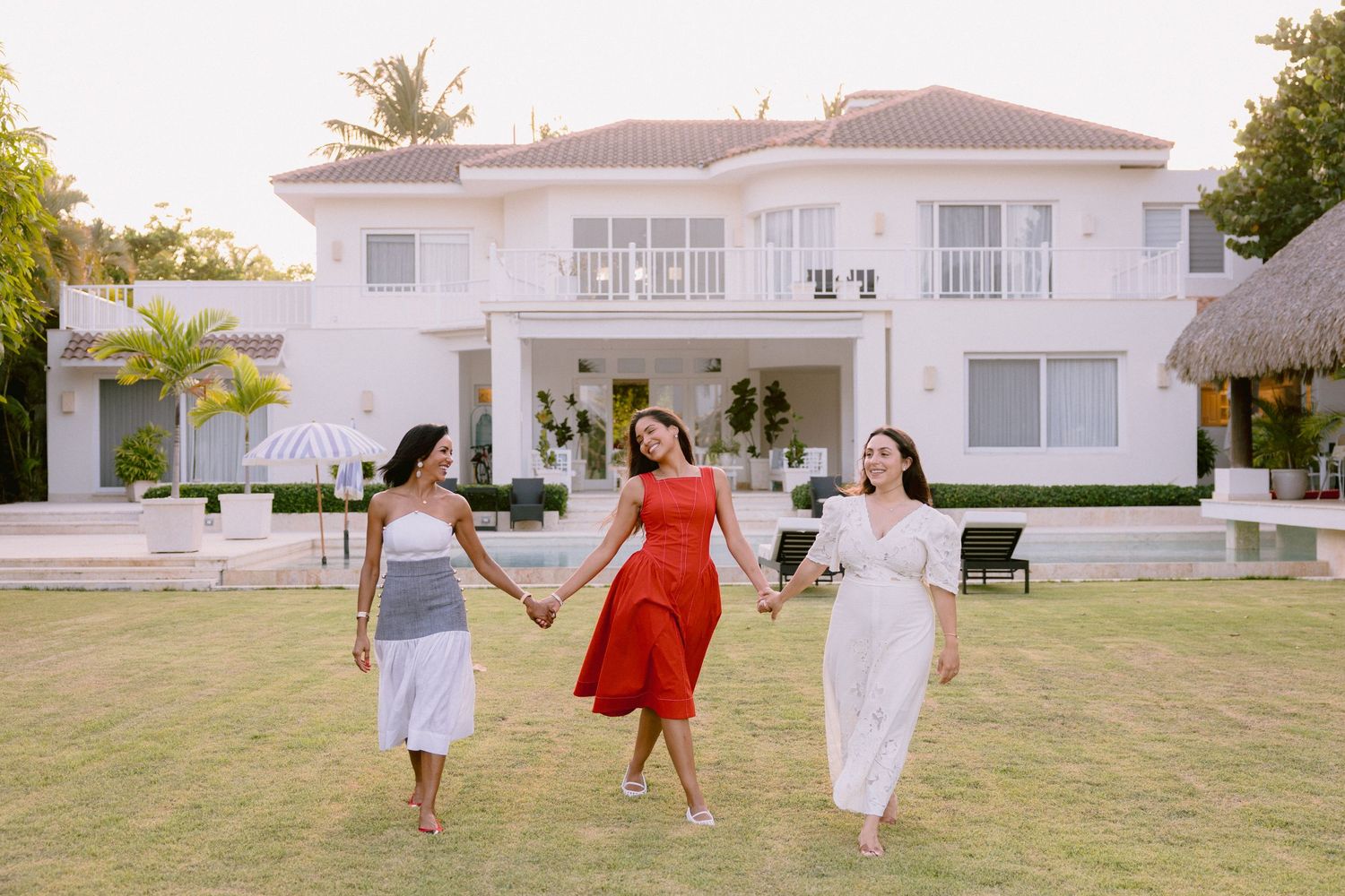 Three women in colorful dresses hold hands and walk on green lawn in front of elegant white villa.
