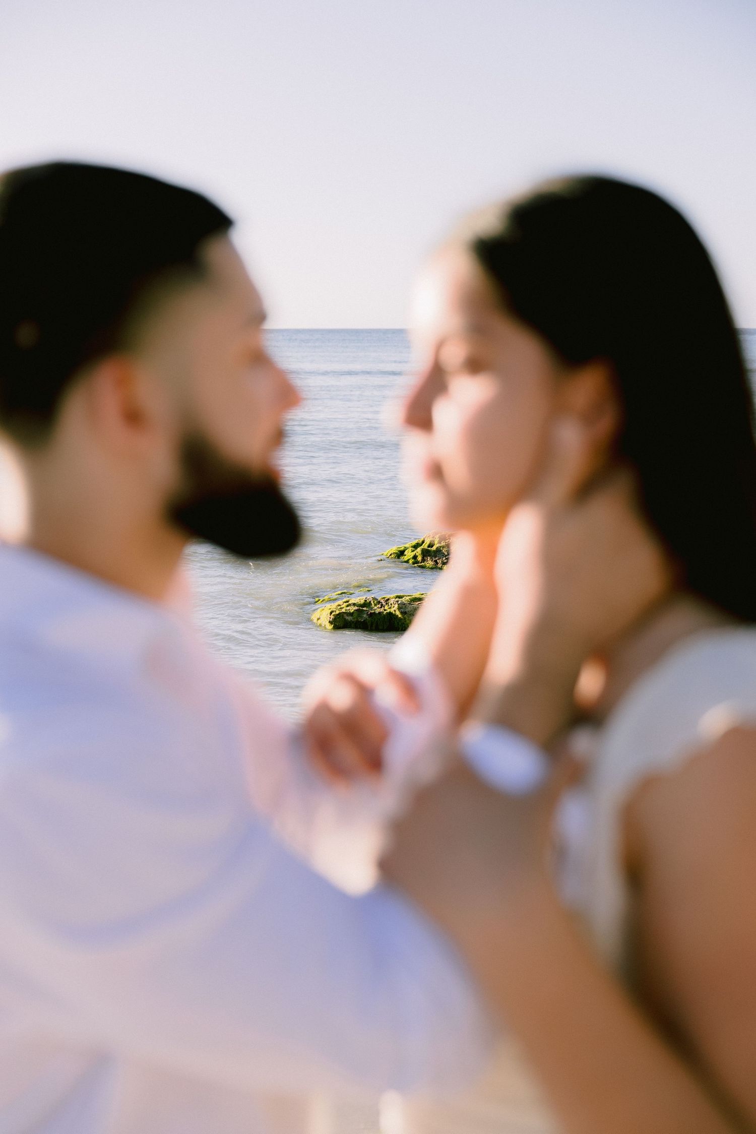 A couple shares a romantic moment by the sea, the bearded man gently touching the woman's face near rocky shores.