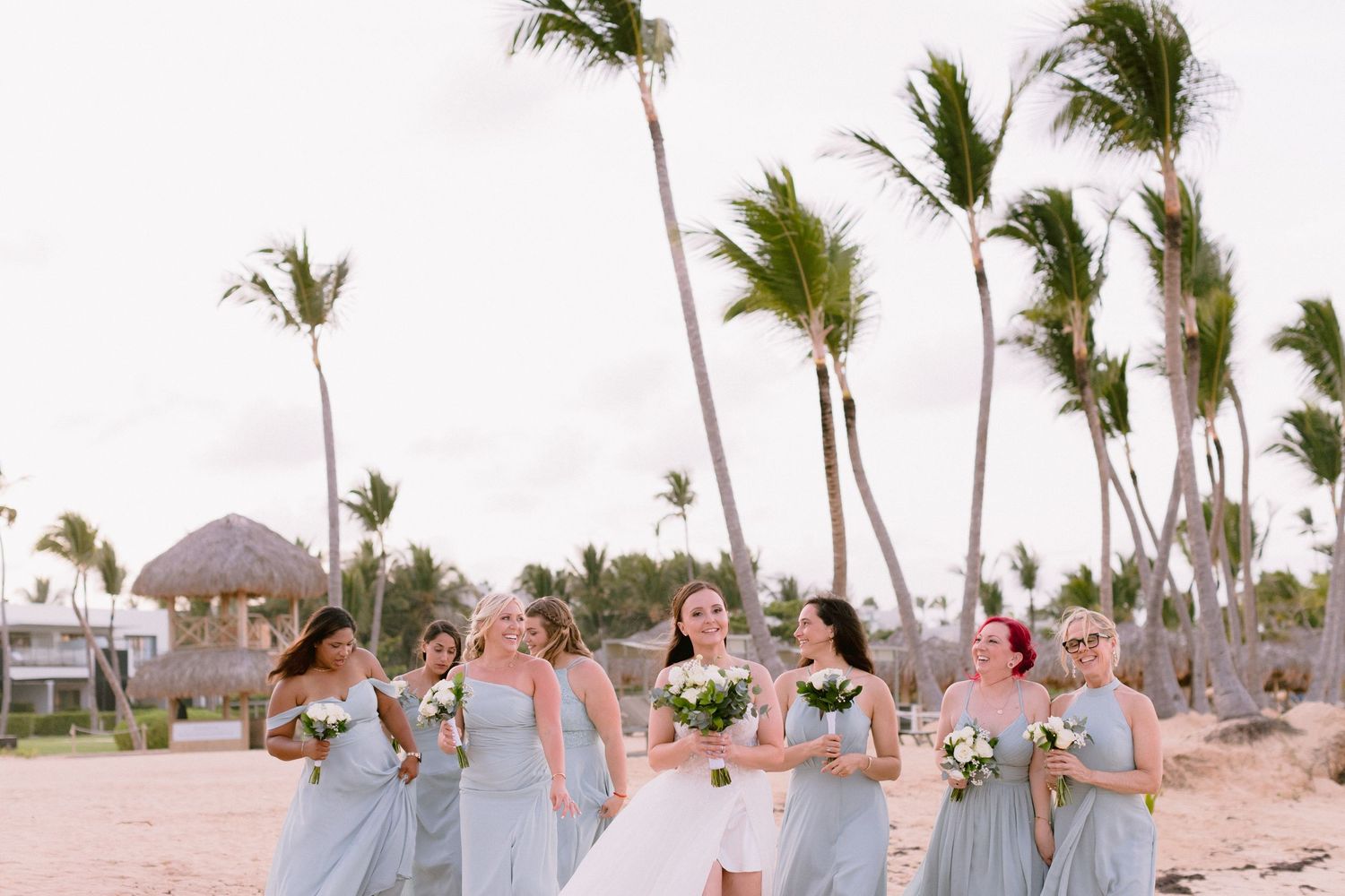 Bride and bridesmaids in light blue dresses laughing on a tropical beach with palm trees swaying in the wind.