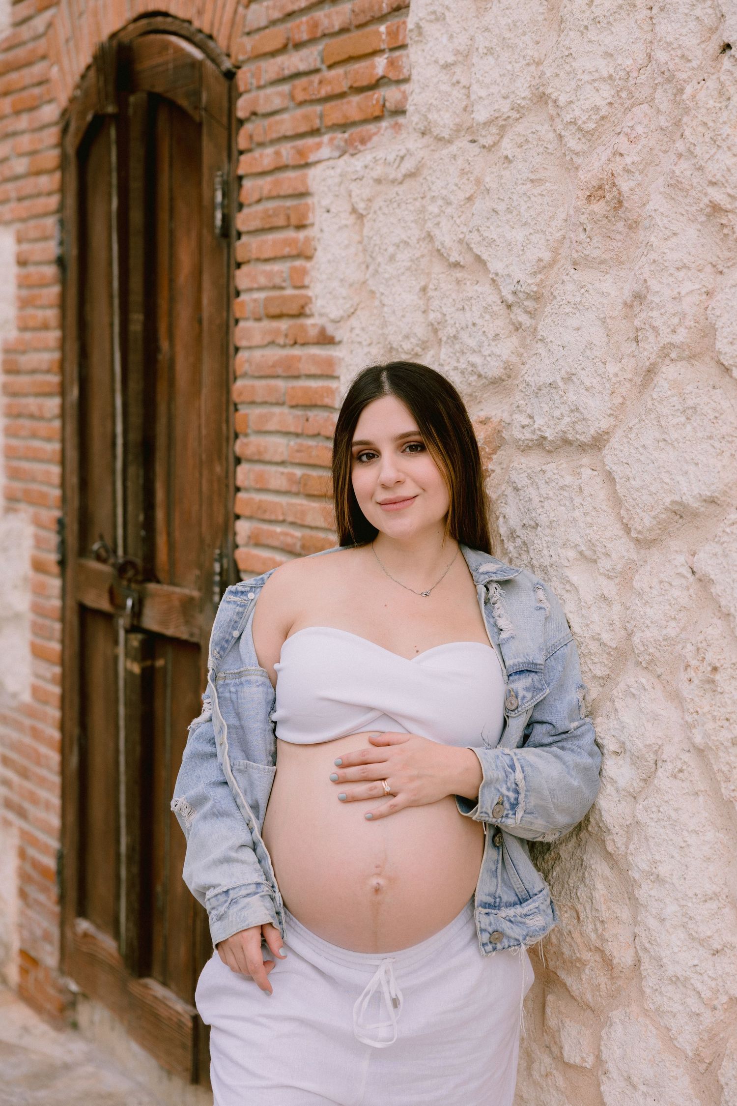 Maternity photo against rustic brick wall wearing white top and denim jacket in warm natural light.