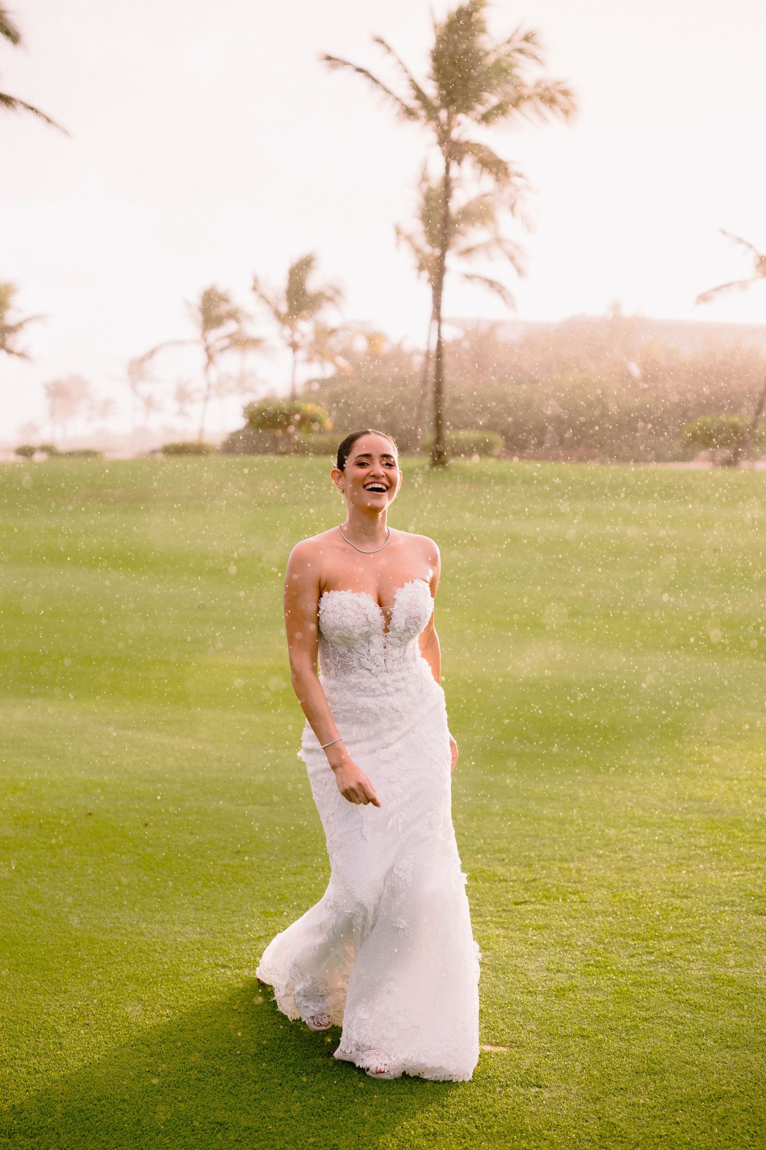 A person in a white wedding dress runs joyfully across a green lawn with palm trees in the background.