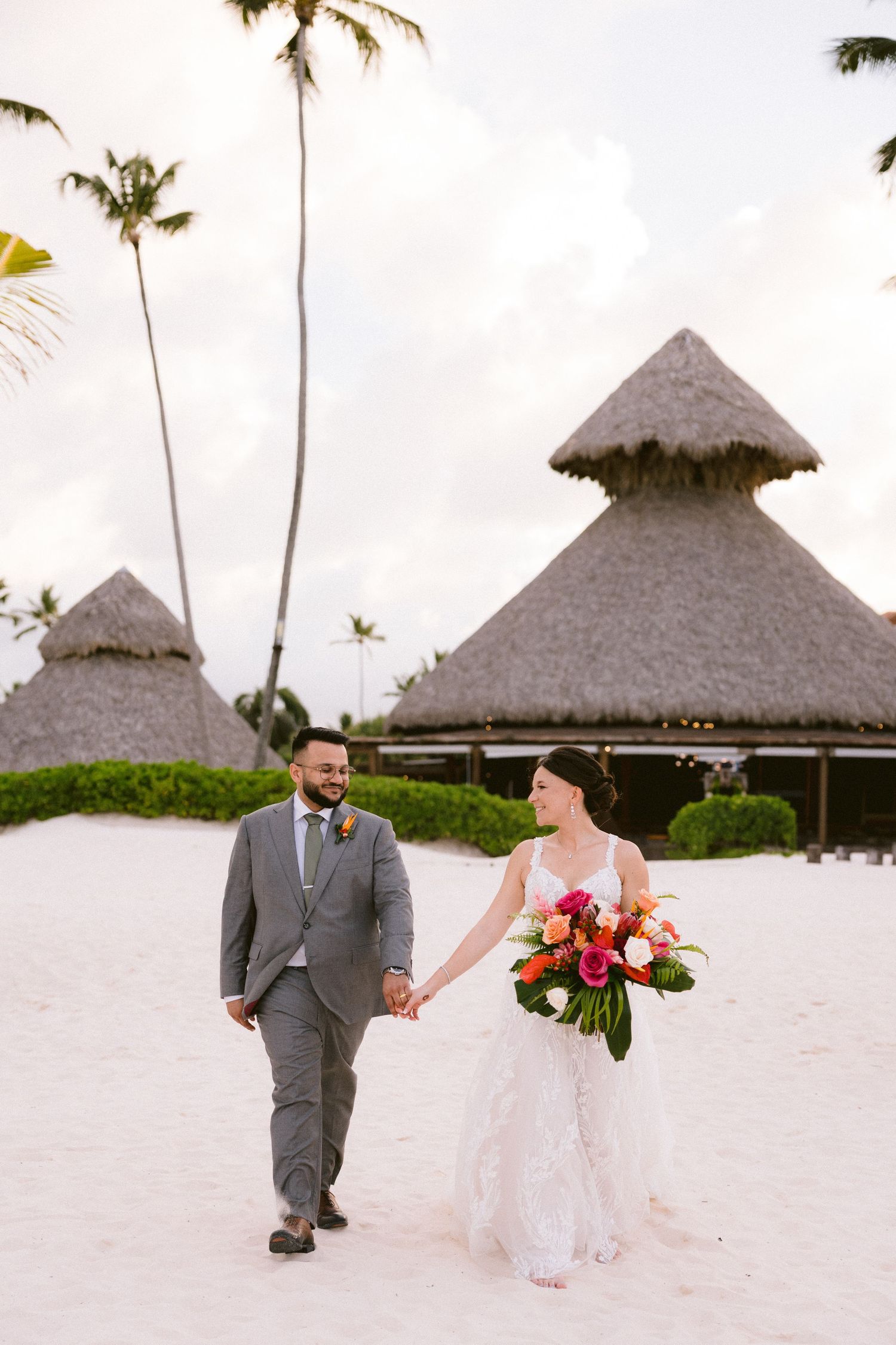 A couple walks hand in hand on a tropical beach with thatched-roof huts and palm trees in the background.