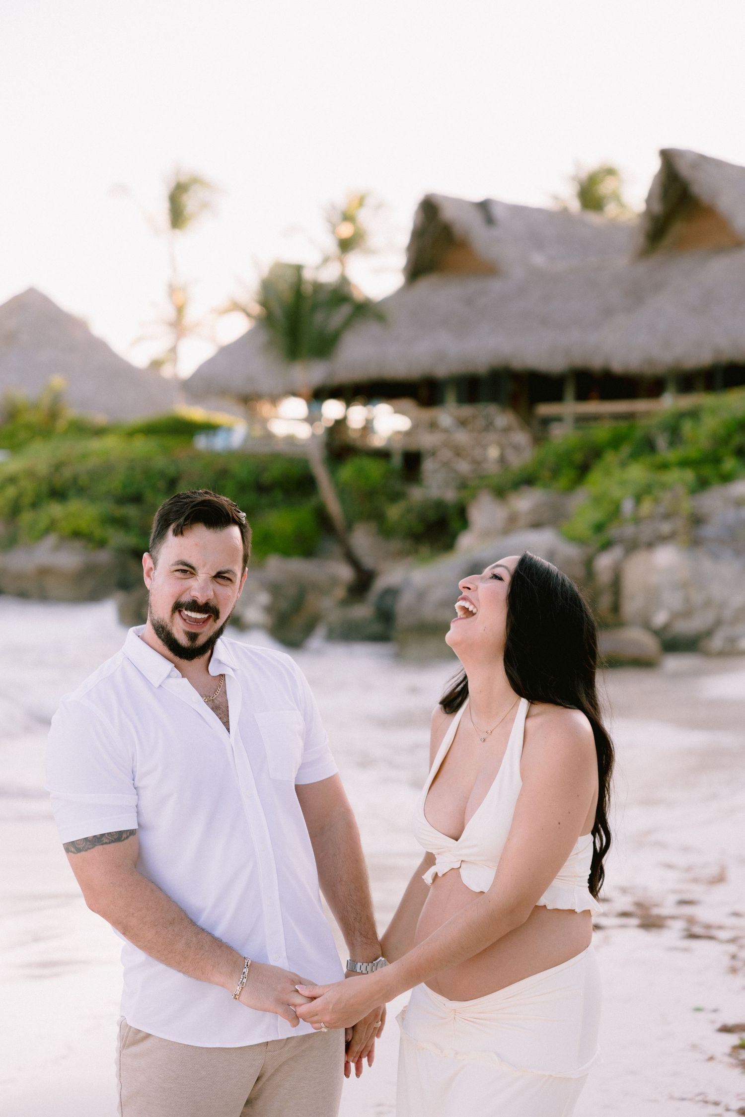 A laughing couple in white outfits holds hands on a tropical beach with thatched huts and palm trees at sunset.