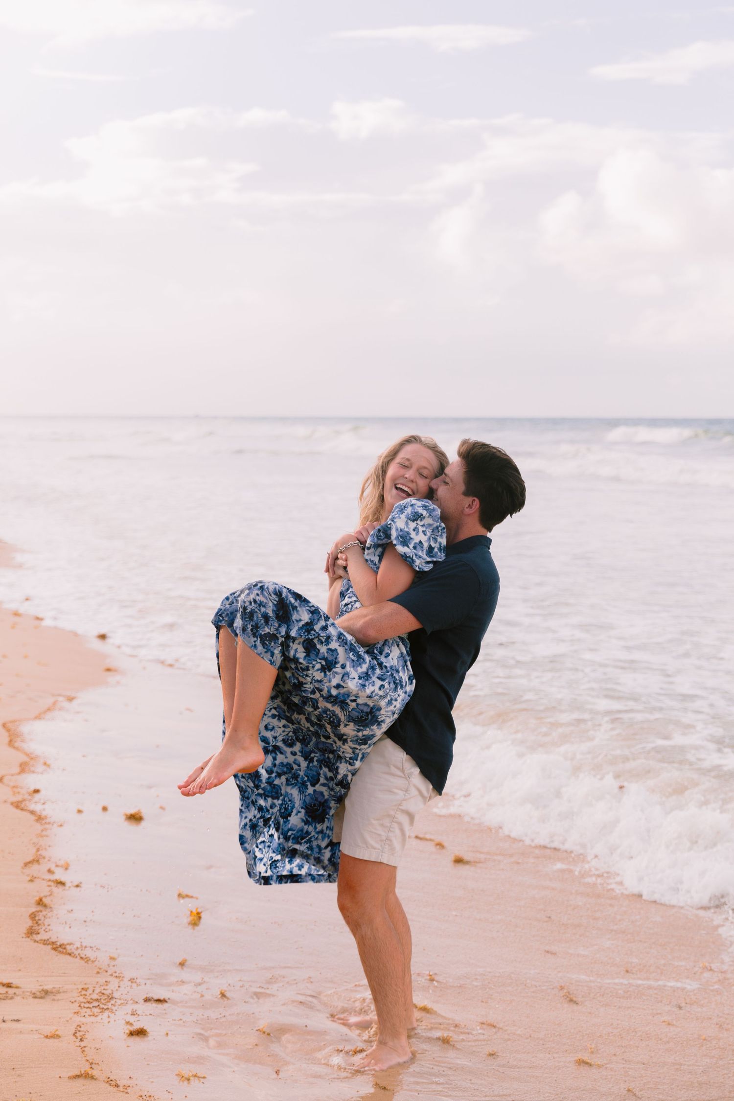 A romantic couple embraces and plays on a sandy beach with ocean waves in the background.