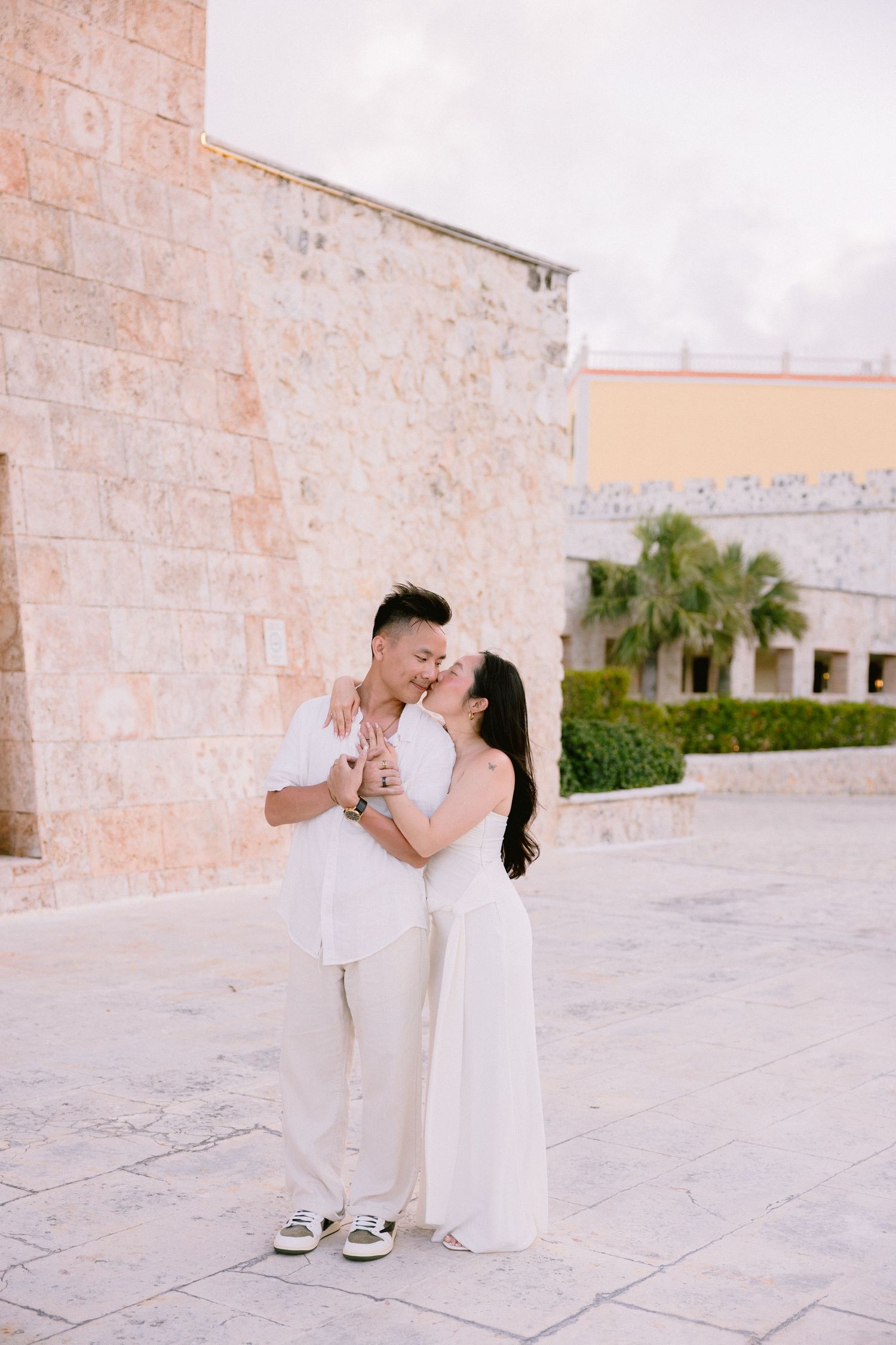 Couple in matching white outfits sharing a romantic kiss near a stone building in a sunny outdoor plaza.
